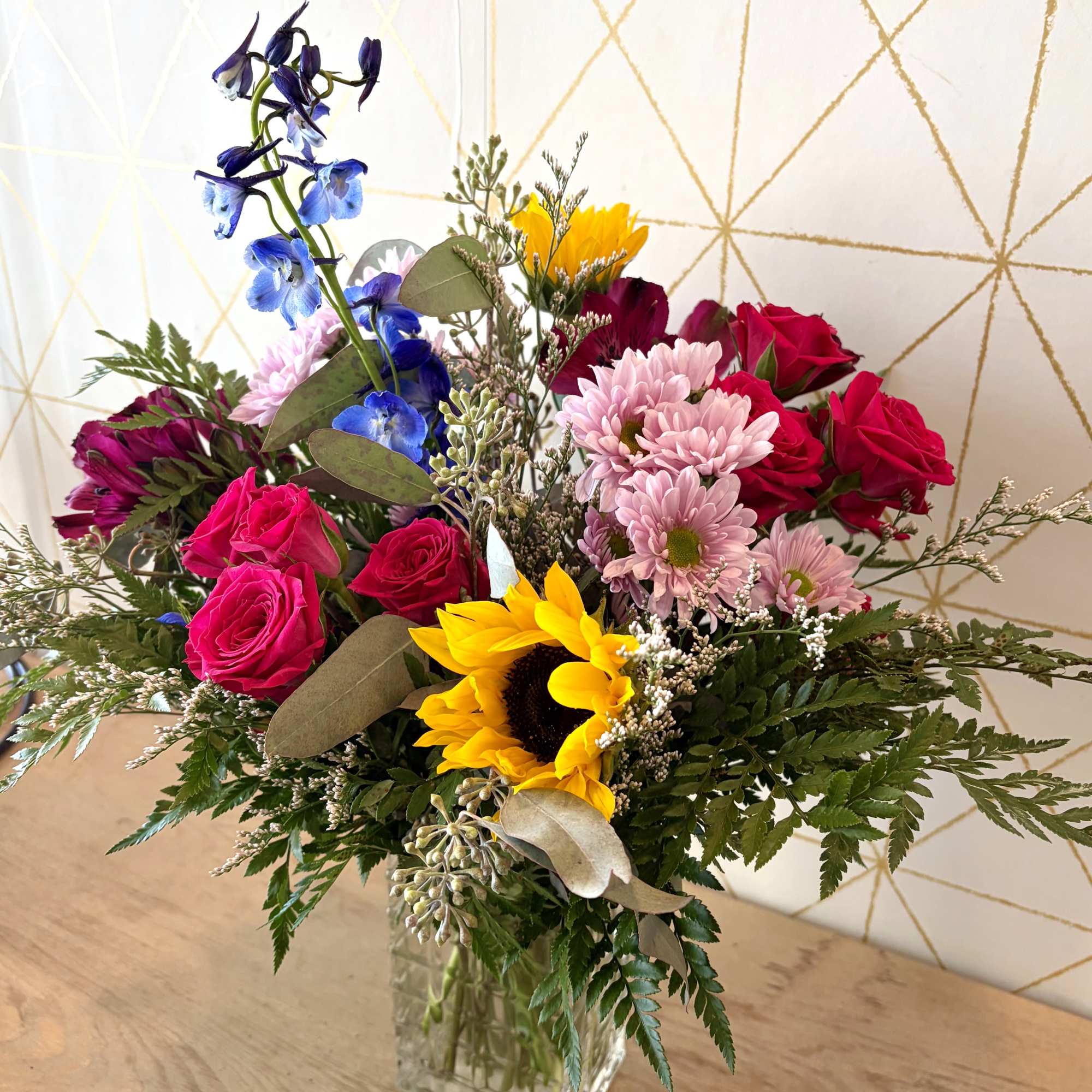 Mixed bouquet with roses, sunflowers, blue delphinium, and pink daisies in a glass vase