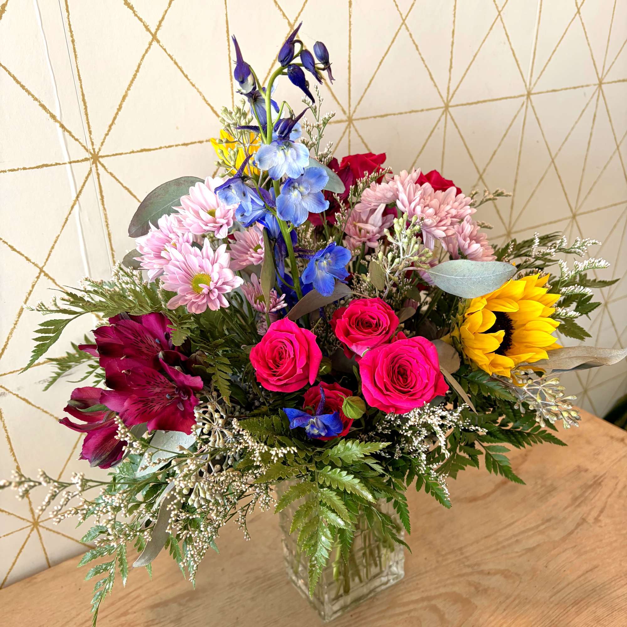 Mixed bouquet with roses, sunflowers, daisies, and blue flowers in a glass vase