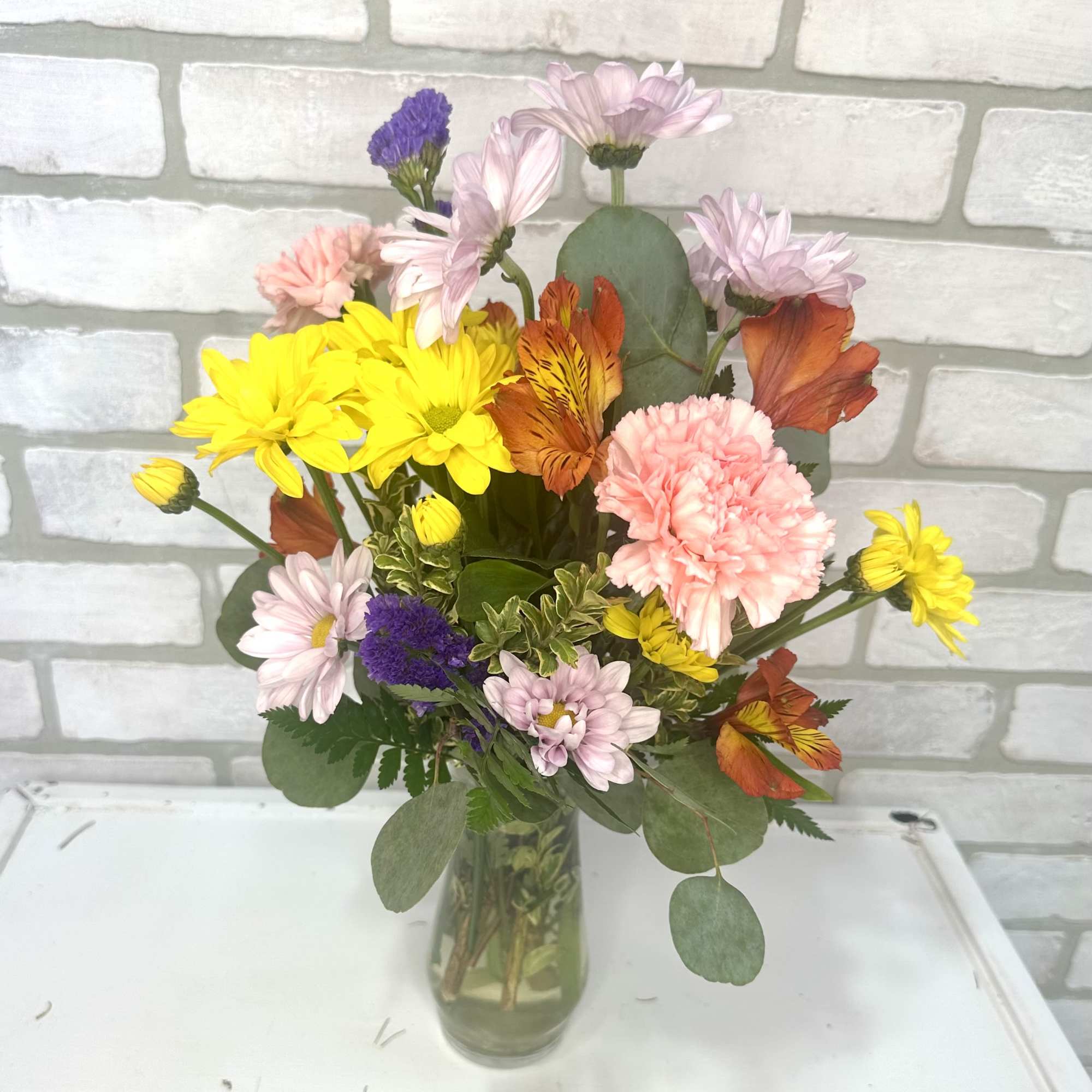 Mixed bouquet of yellow daisies, pink carnations, and orange blooms in a clear glass vase