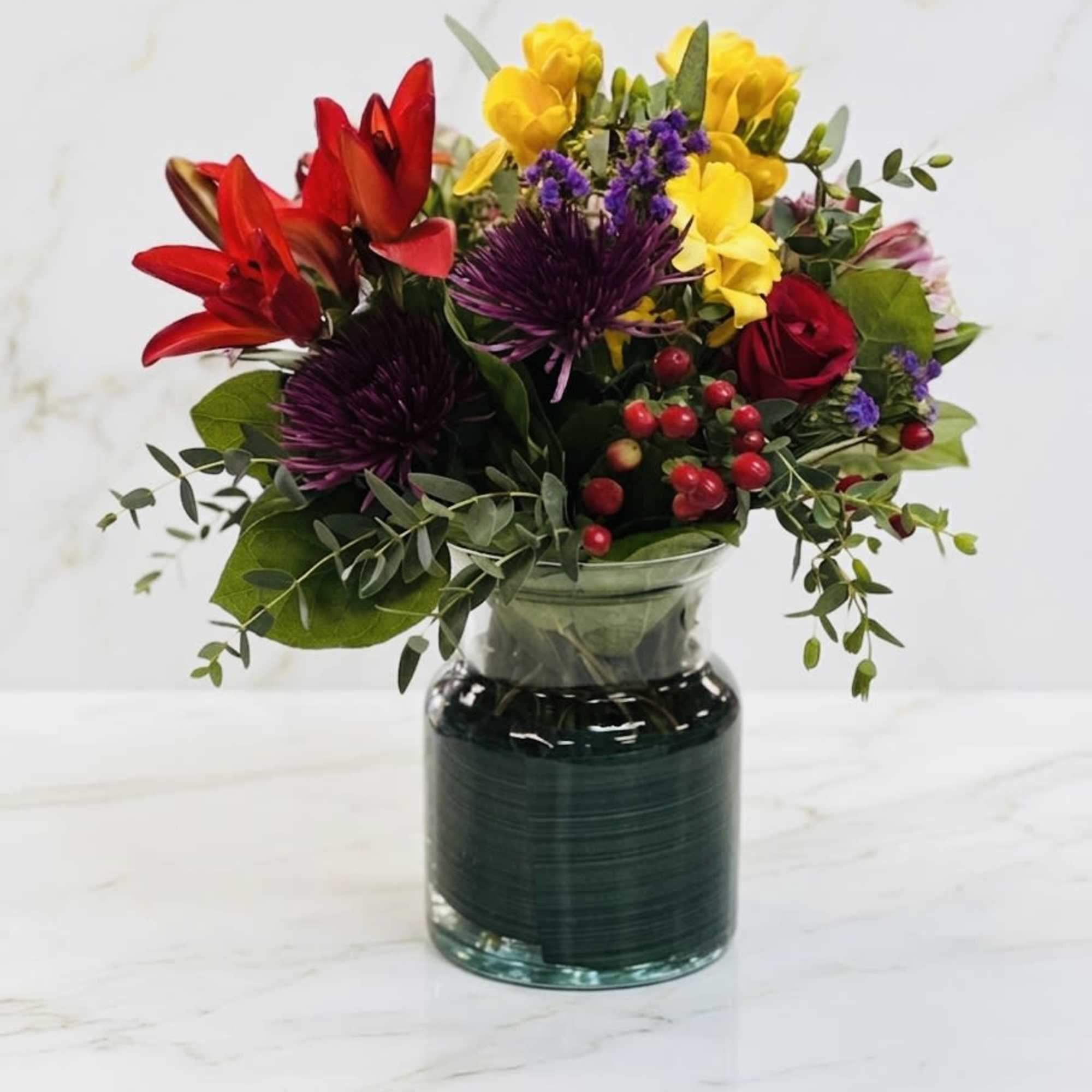Bright mixed bouquet with red lilies, yellow blooms, purple mums and red roses in a clear glass vase