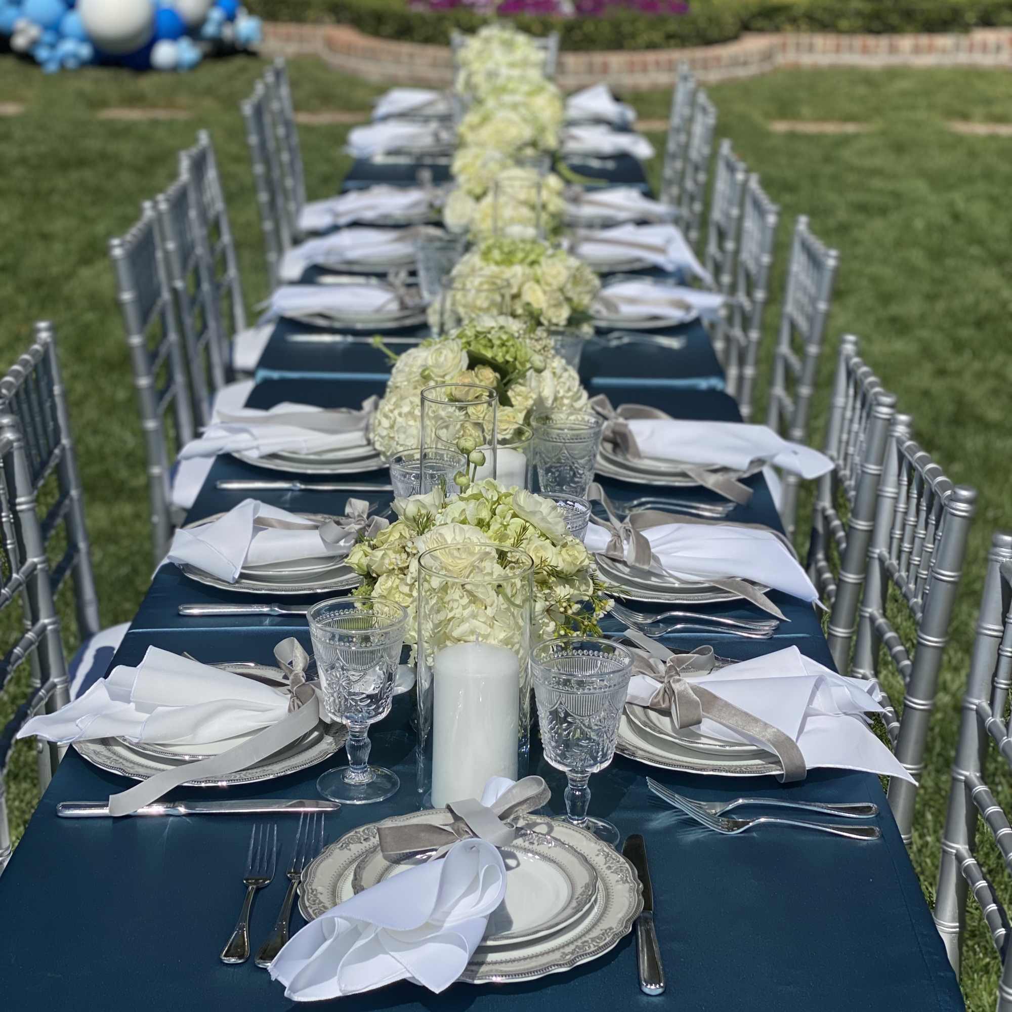Long outdoor wedding table with white floral centerpieces and silver place settings
