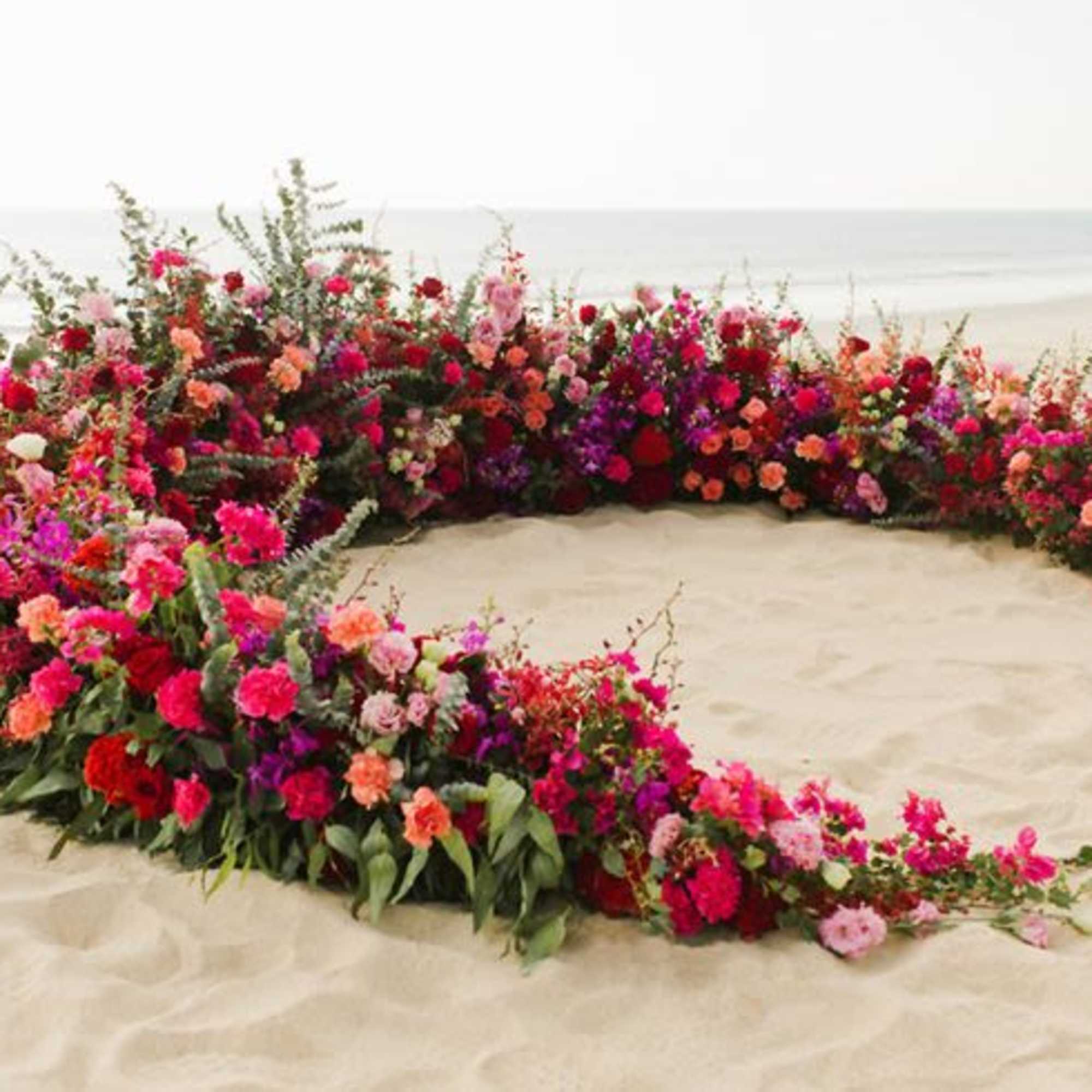 Large crescent floral arrangement on sand with pink, red, and orange blooms
