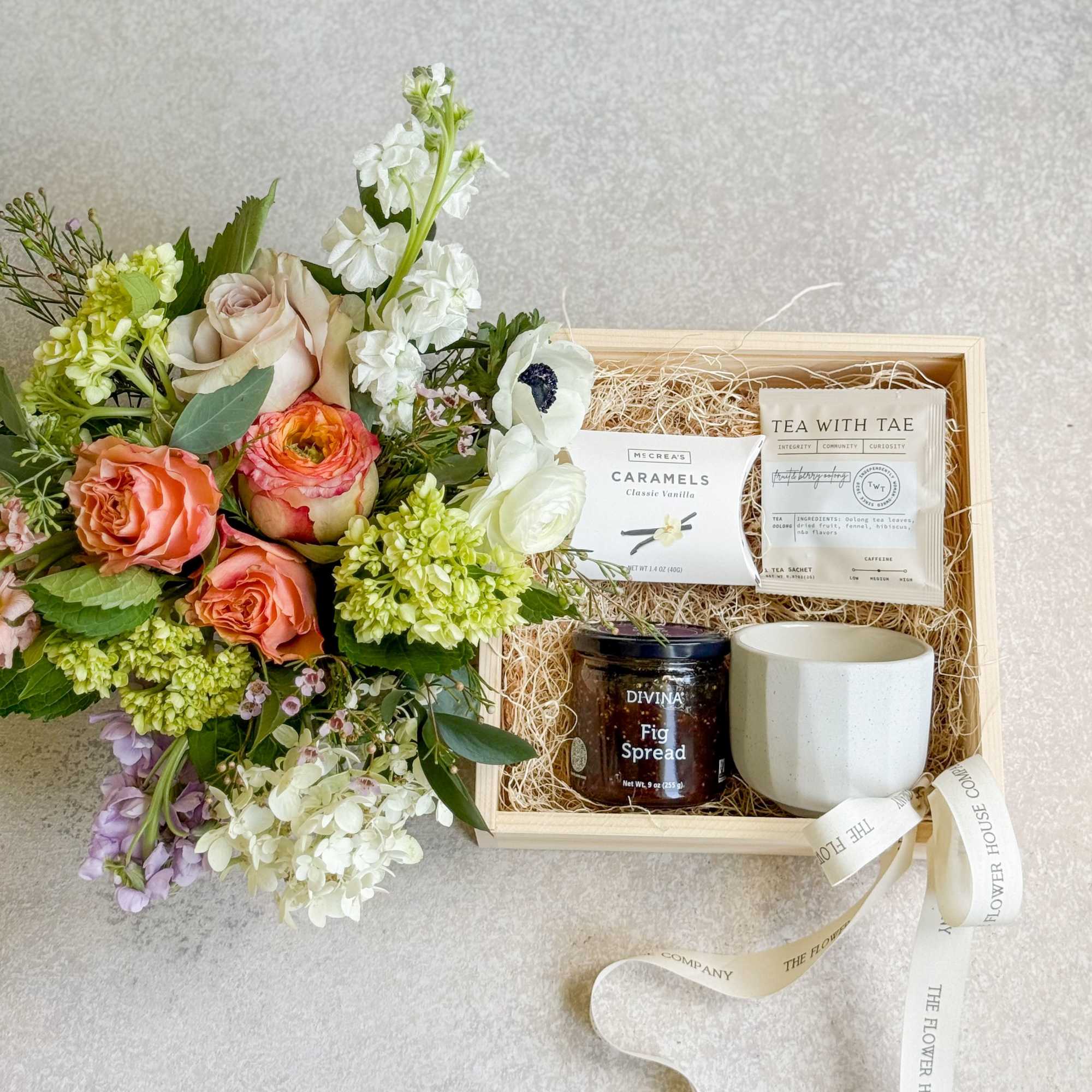 Floral bouquet beside a gift box with tea, caramels, fig spread, and a mug