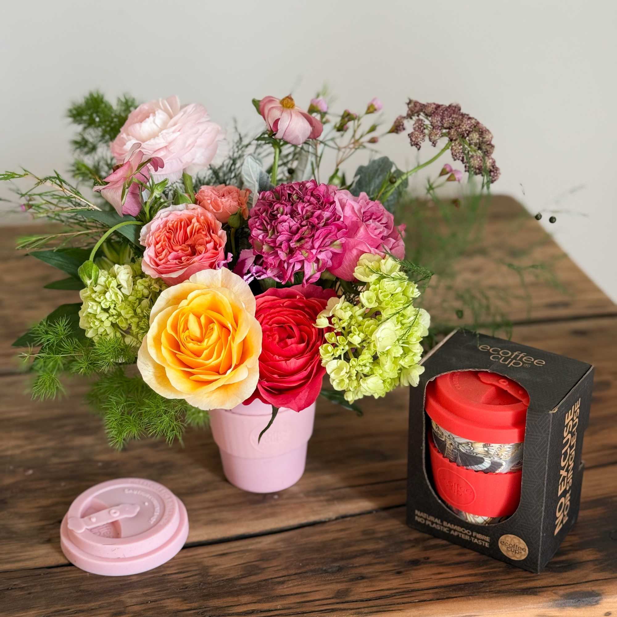 Mixed bouquet in a pink cup with a red travel mug and lid beside it