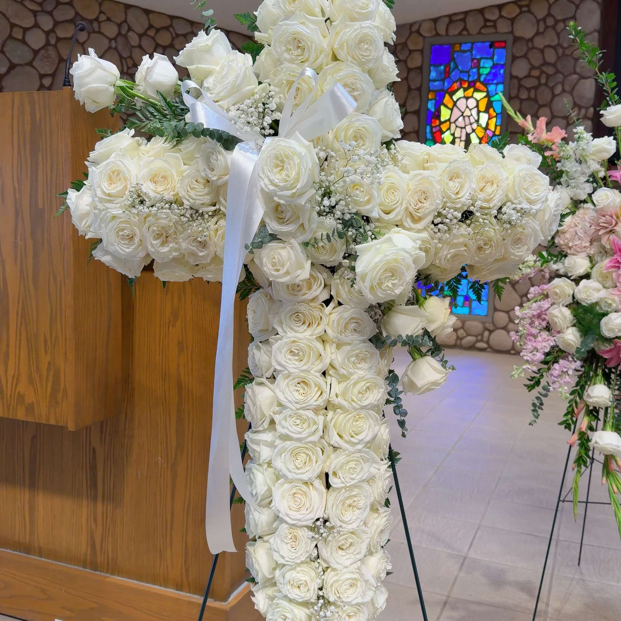 White rose cross-shaped funeral spray on a stand with a white ribbon