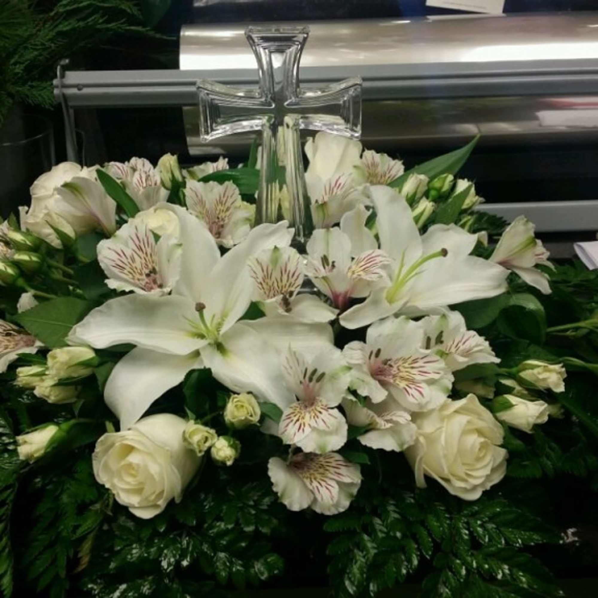 White floral casket spray with lilies, roses, and alstroemeria around a clear cross