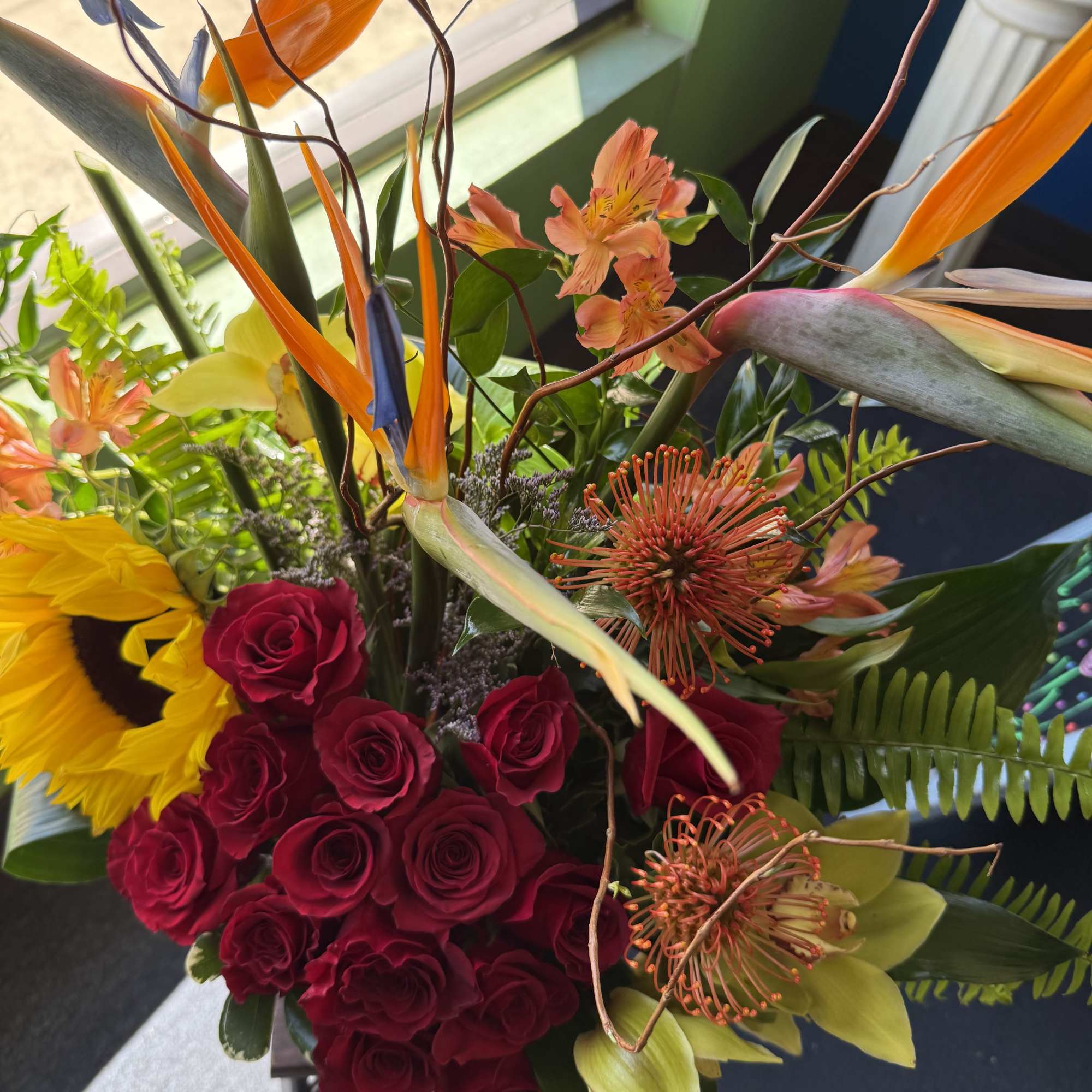Bright tropical arrangement with red roses, sunflower, birds of paradise, protea, and green orchids