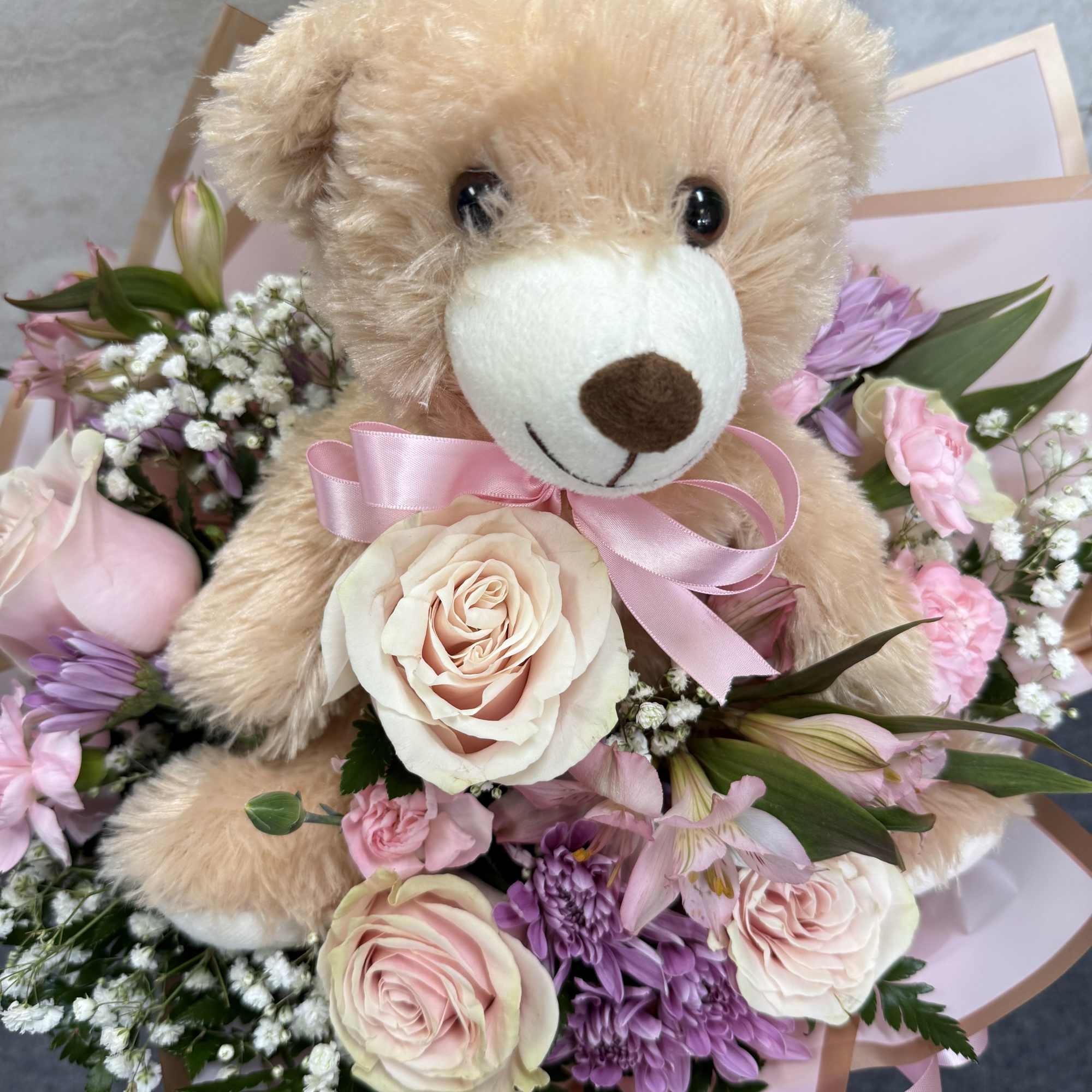 Teddy bear surrounded by pink and purple flowers with a ribbon bow