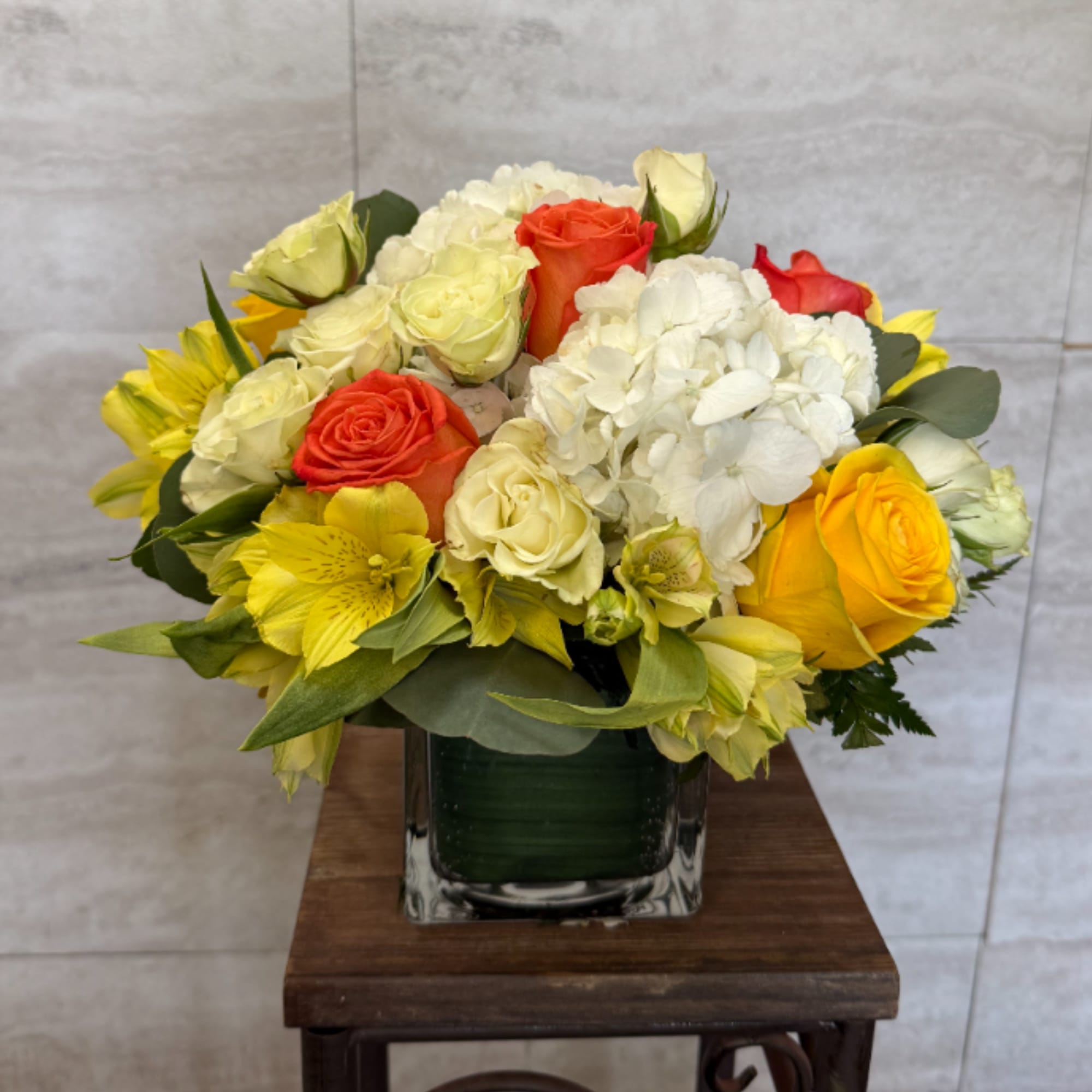Compact arrangement of yellow, white, and orange flowers in a clear glass cube vase.
