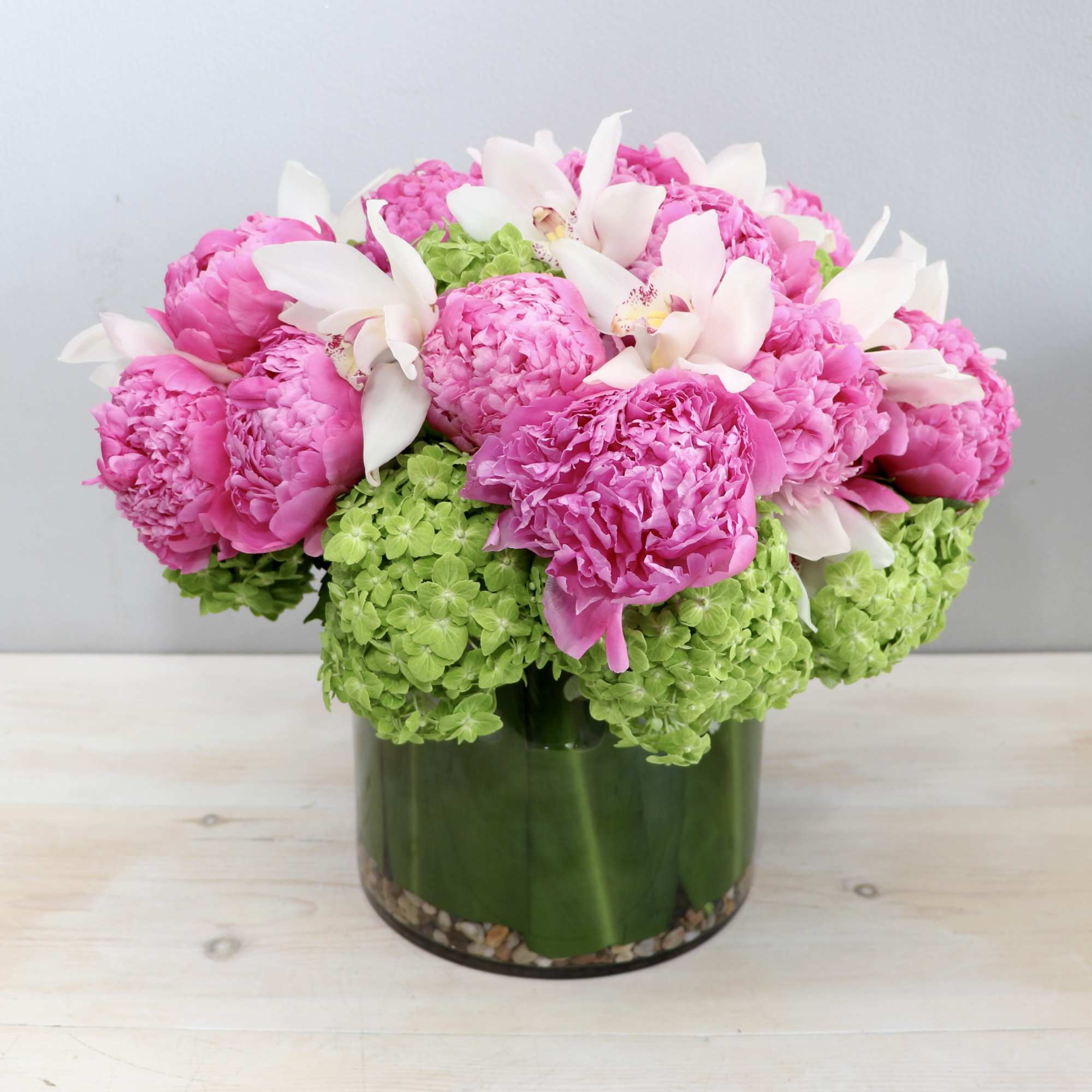 Low arrangement of bright pink peonies, white orchids, and green hydrangeas in a round glass vase