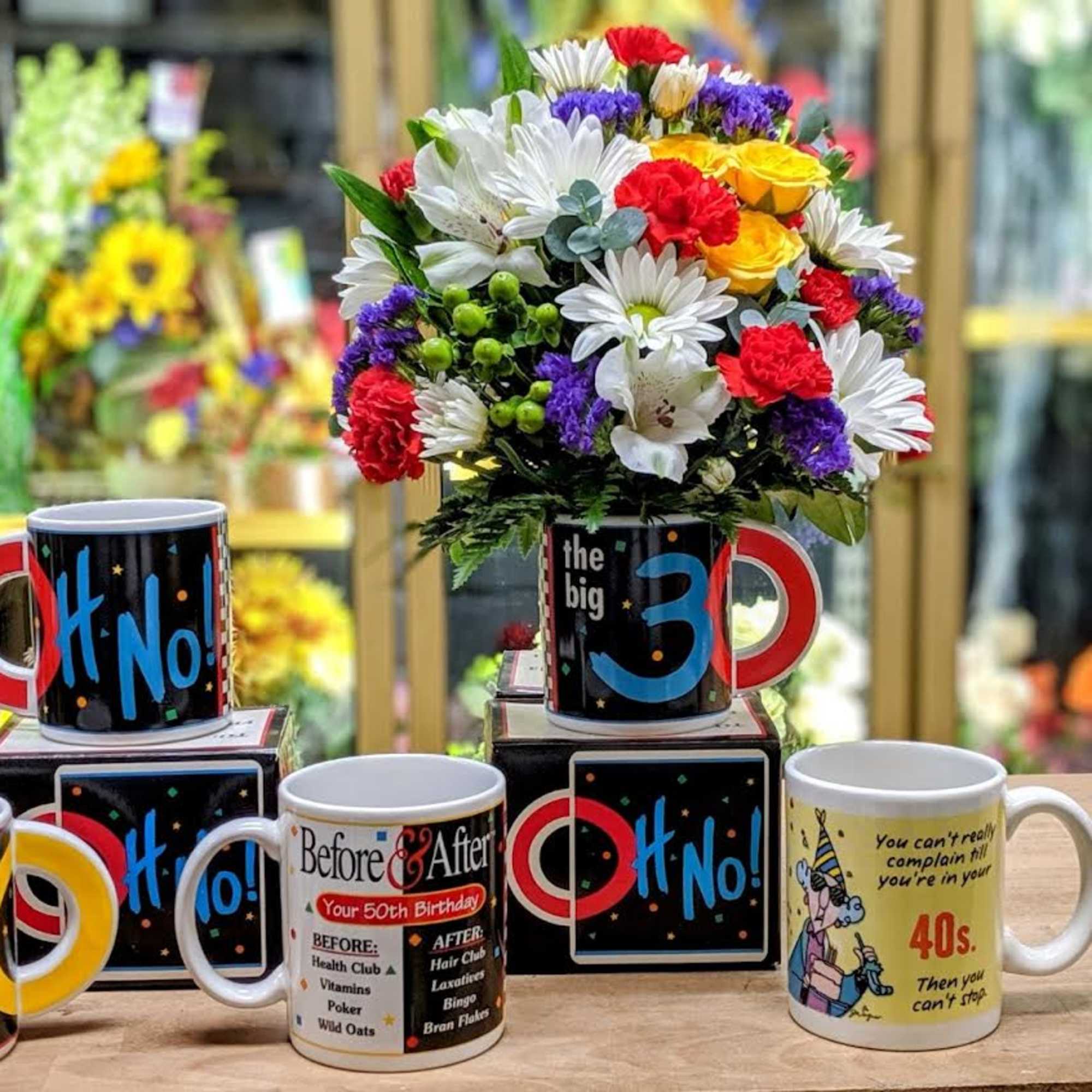 Colorful mixed flower bouquet in a mug-shaped container with novelty birthday mugs nearby