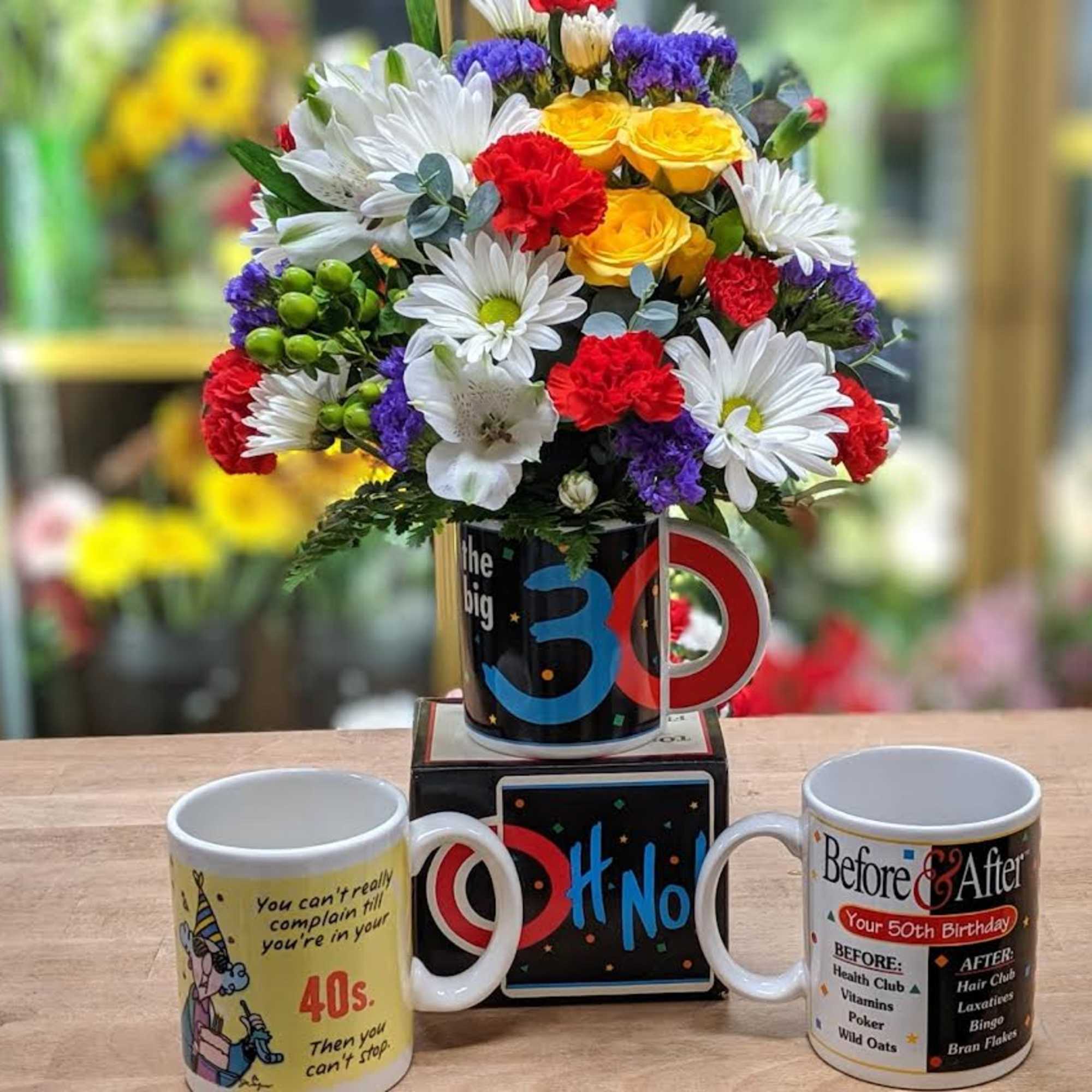 Colorful mixed flower arrangement in a novelty mug with two birthday mugs beside it