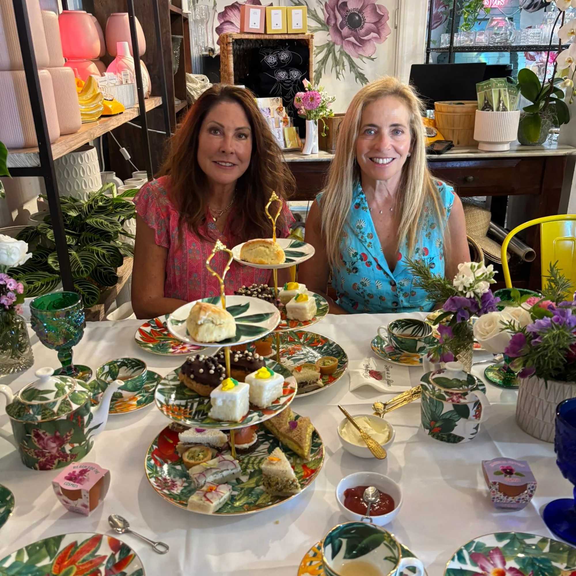 Two women sit at a table with floral dishes, tea, and pastries in a flower shop.