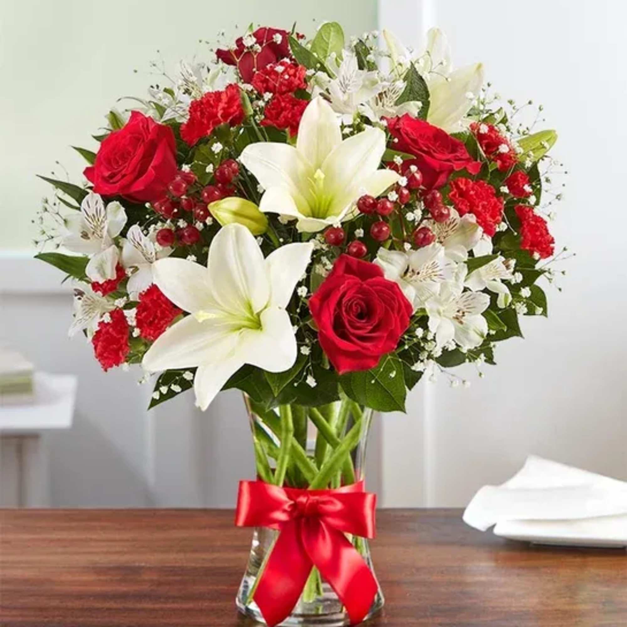 Red roses, white lilies, and red carnations in a clear glass vase with a red ribbon bow