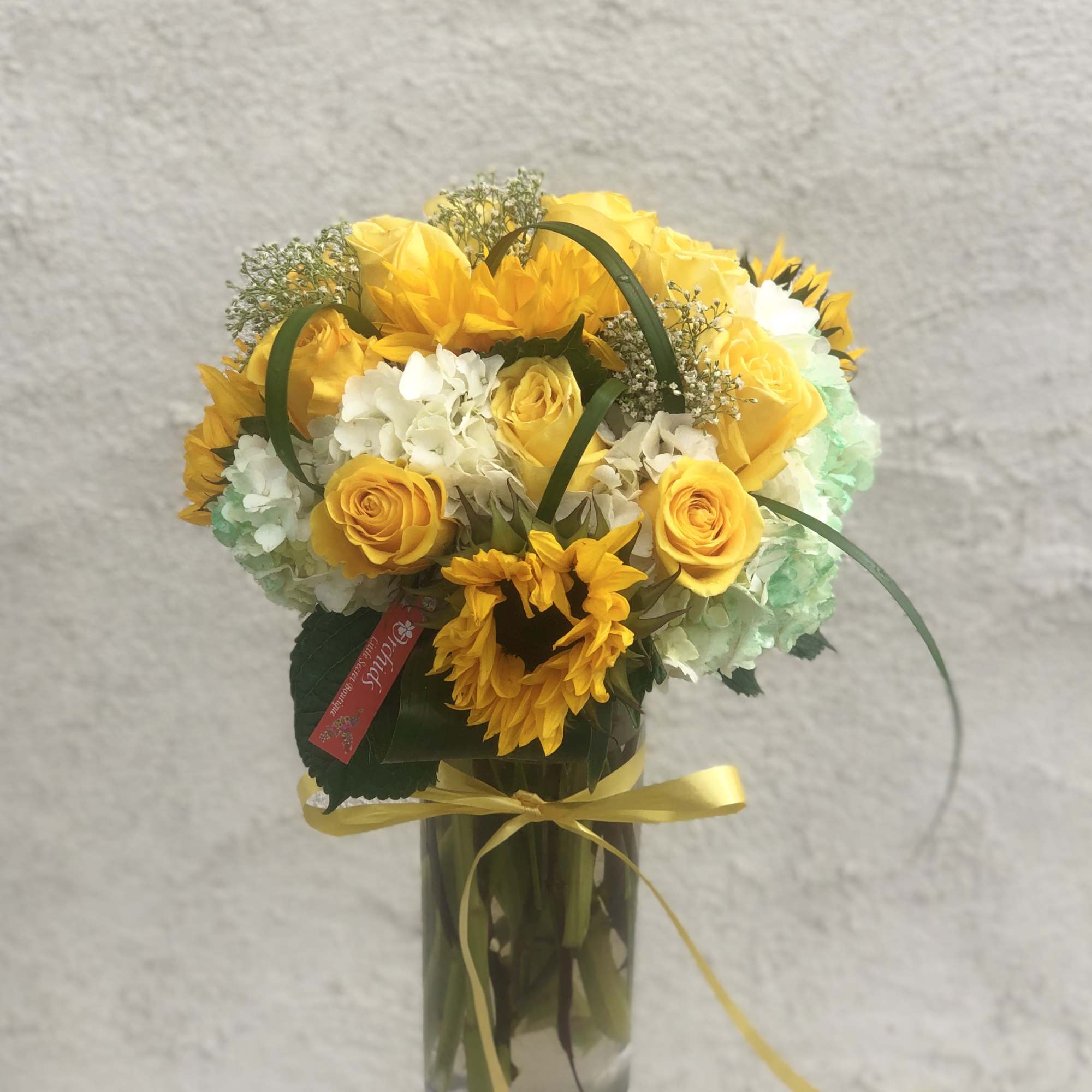 Yellow roses and sunflowers arranged in a clear glass vase with a yellow ribbon