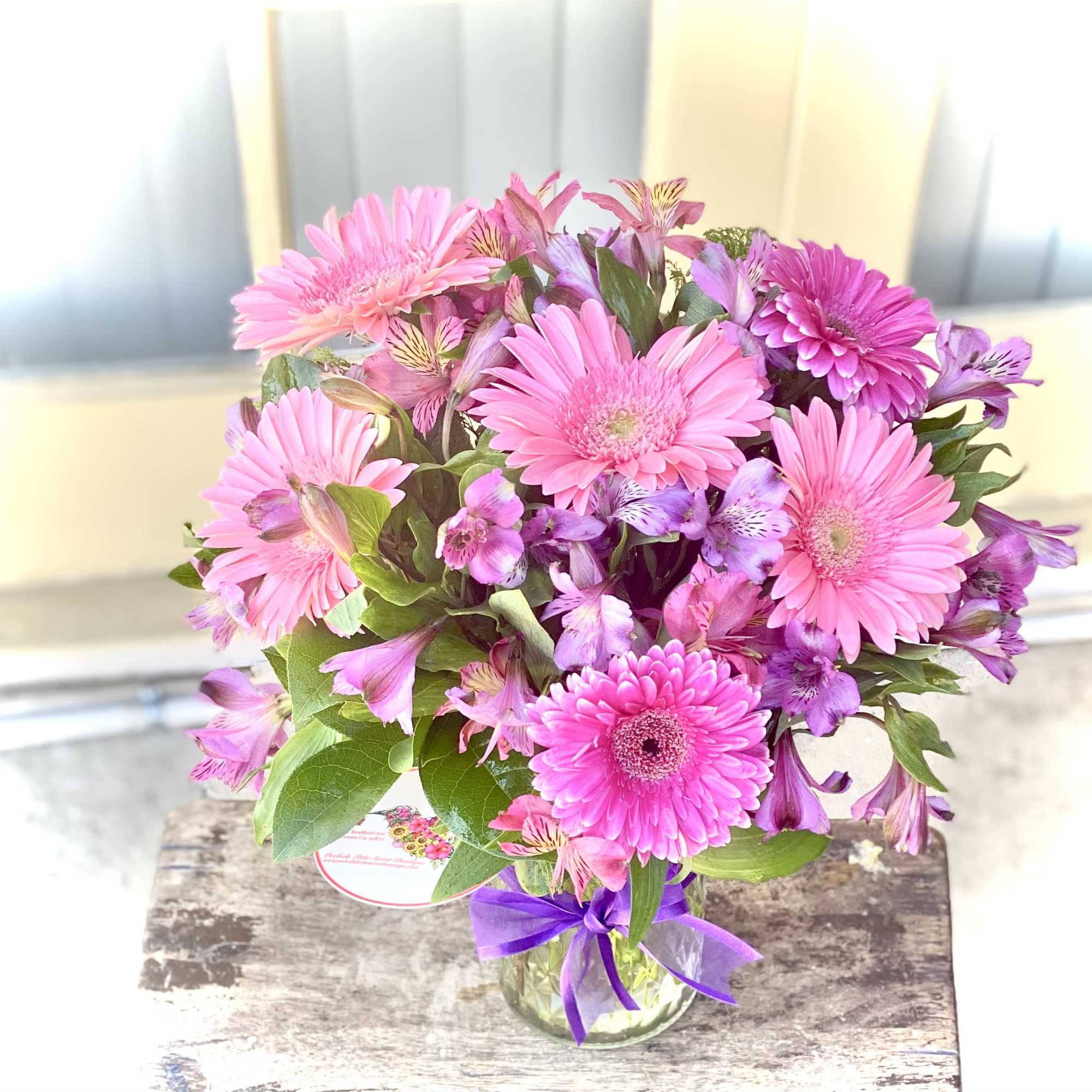 Pink gerbera daisies and purple alstroemeria in a glass vase with a ribbon