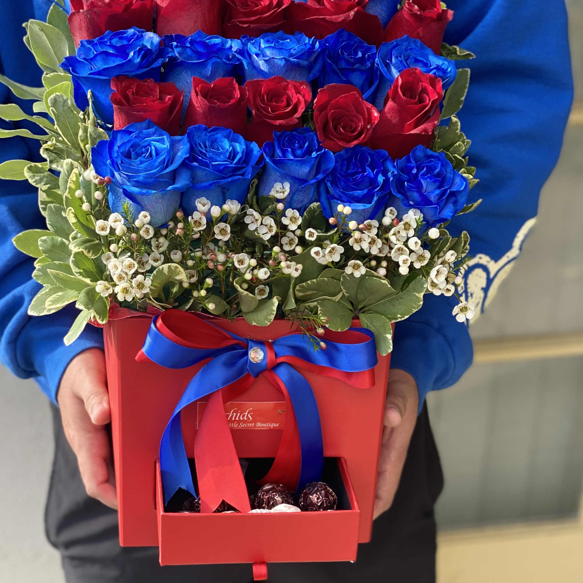 Red and blue roses arranged in a red gift box with a ribbon and chocolates.