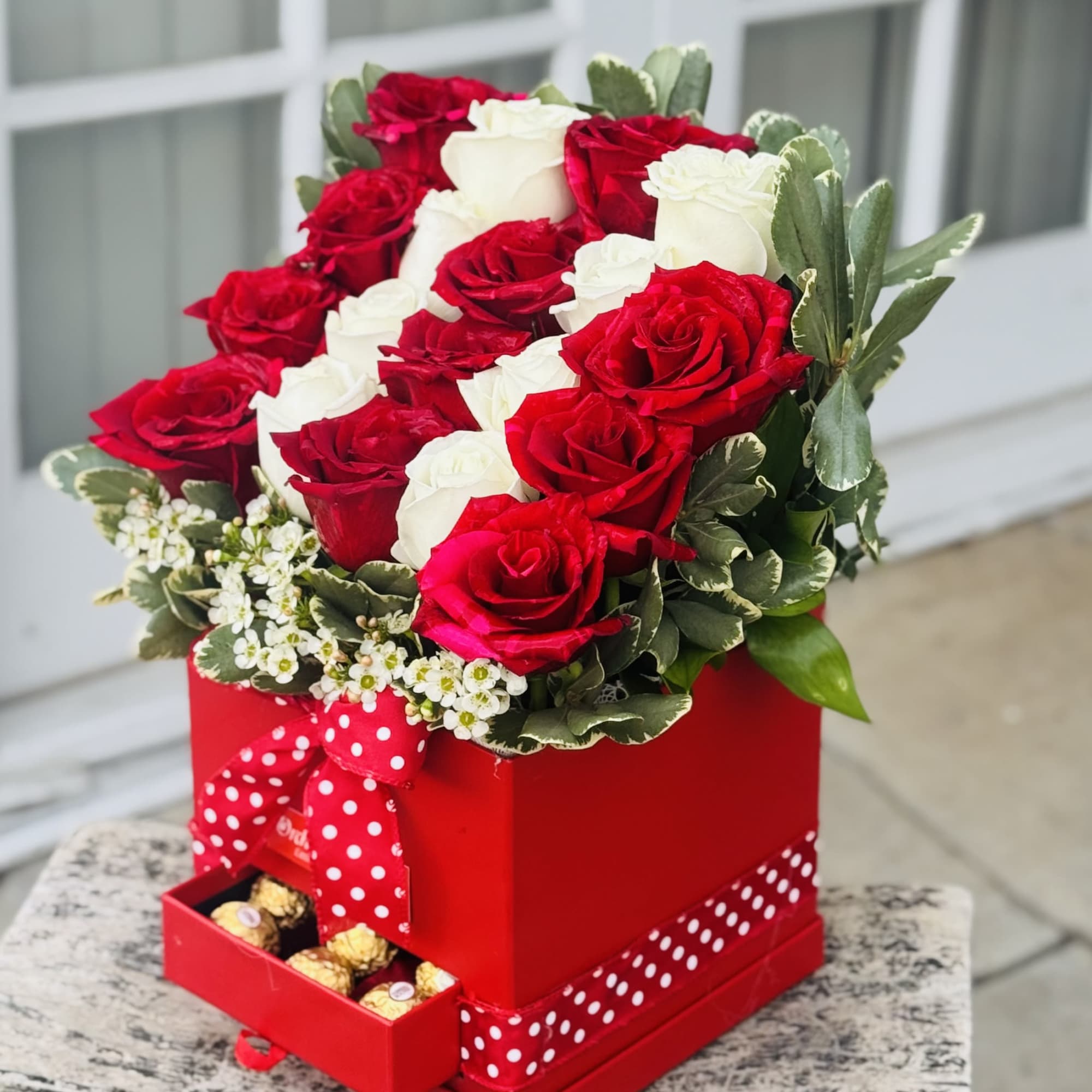 Red and white roses arranged in a red gift box with a ribbon and chocolates