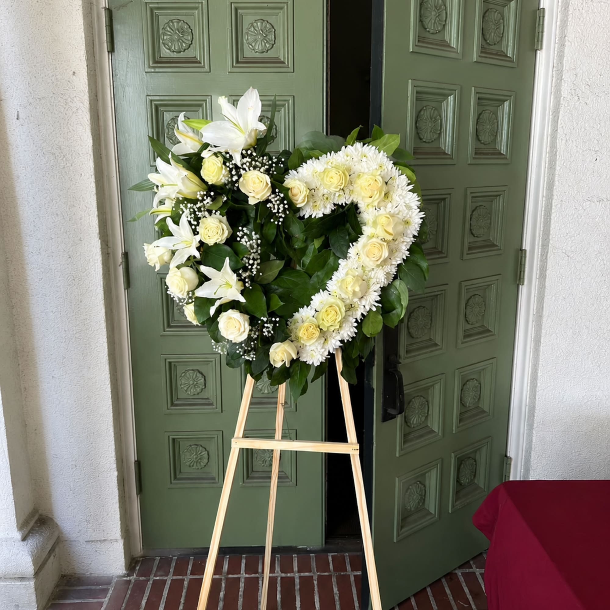 Heart-shaped floral wreath of white lilies, roses, and daisies on a wooden easel