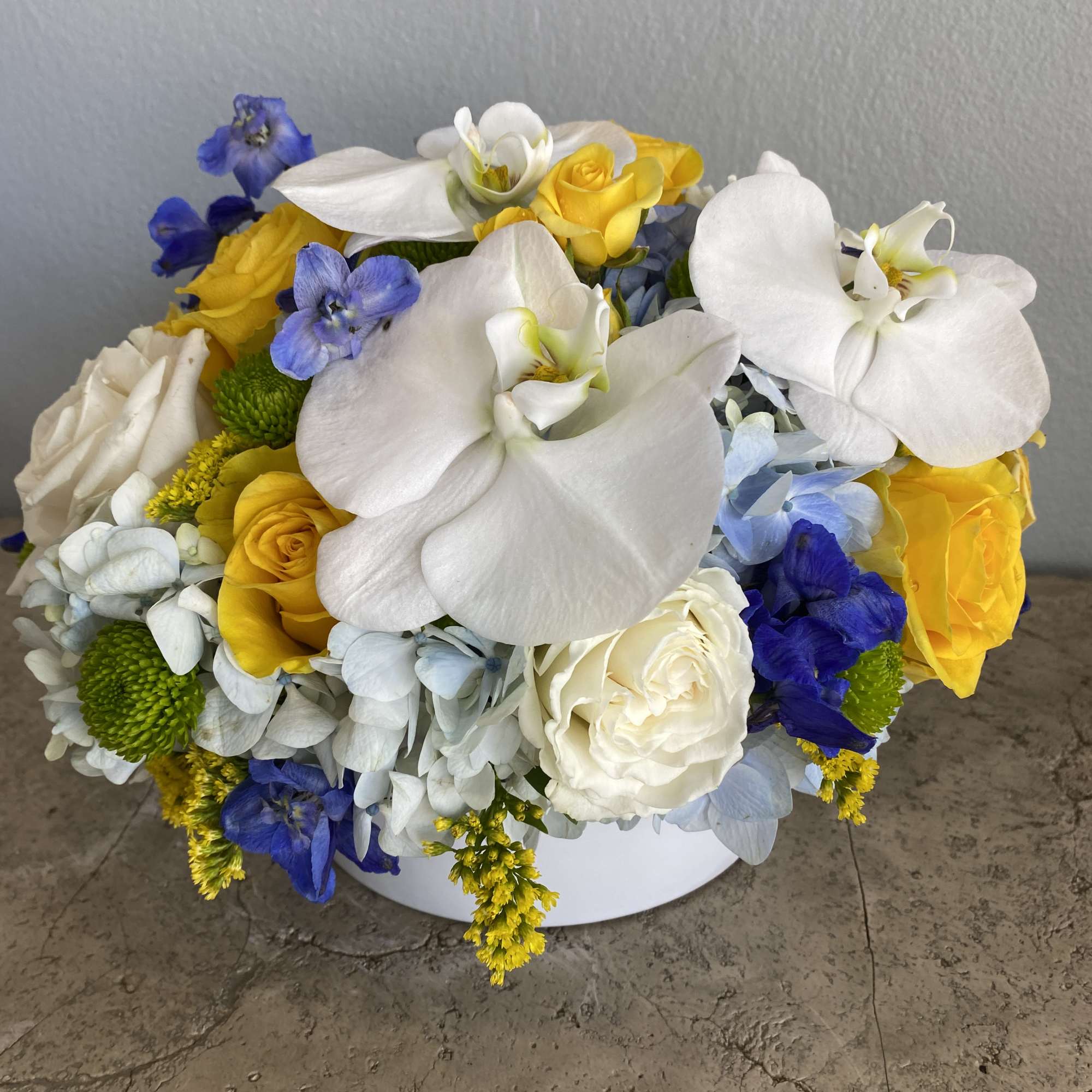 White orchids, yellow roses, and blue flowers in a low white container