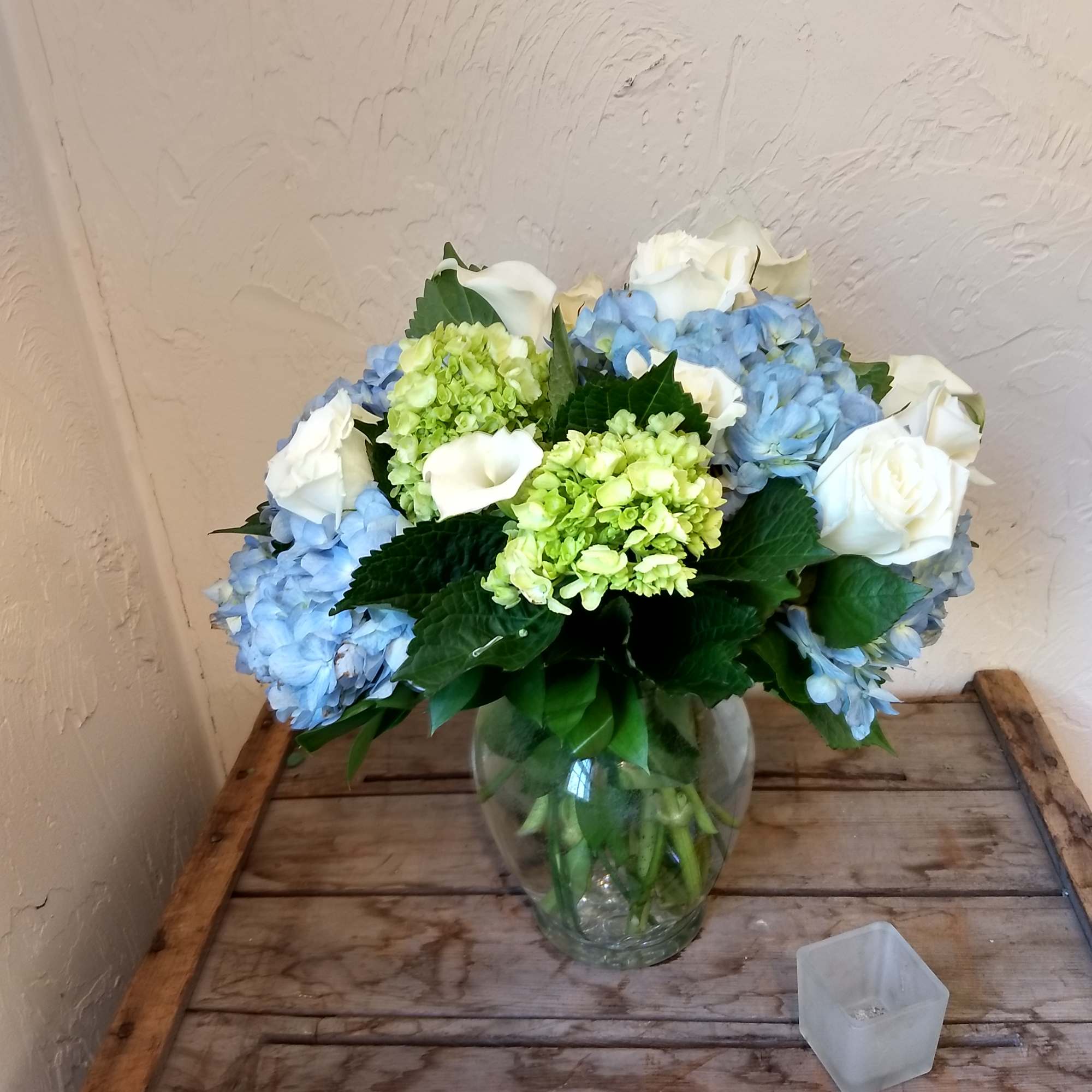 Blue and green hydrangeas with white roses and calla lilies in a clear glass vase on a wooden table