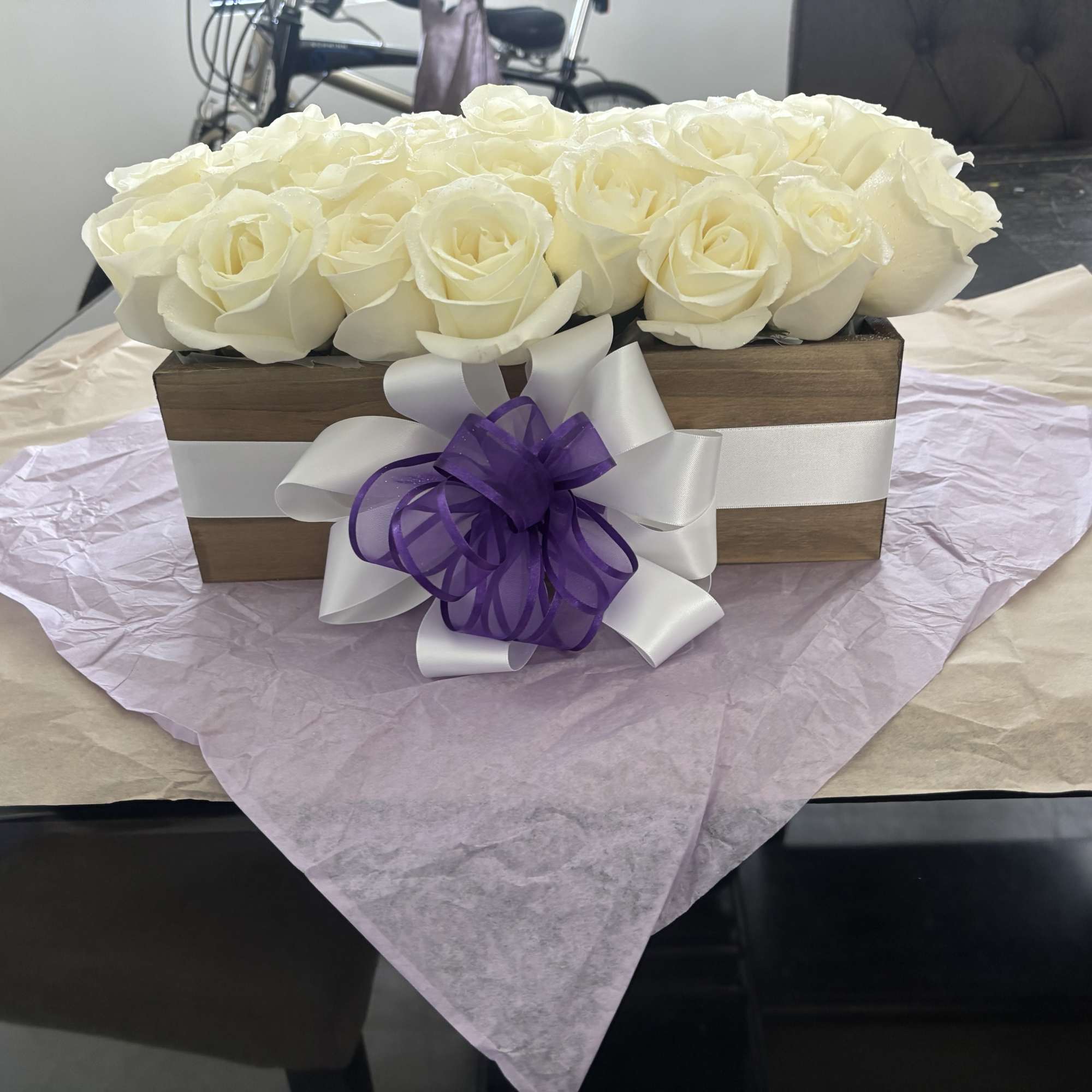White roses arranged in a wooden box with a purple ribbon bow