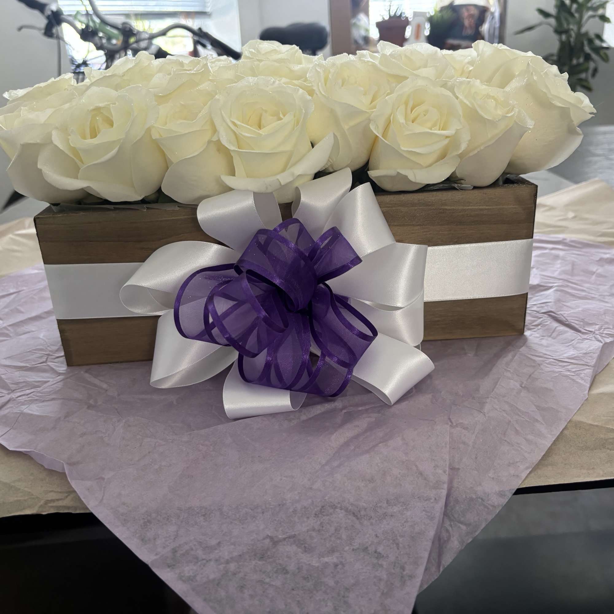 White roses arranged in a wooden box with a white and purple ribbon bow