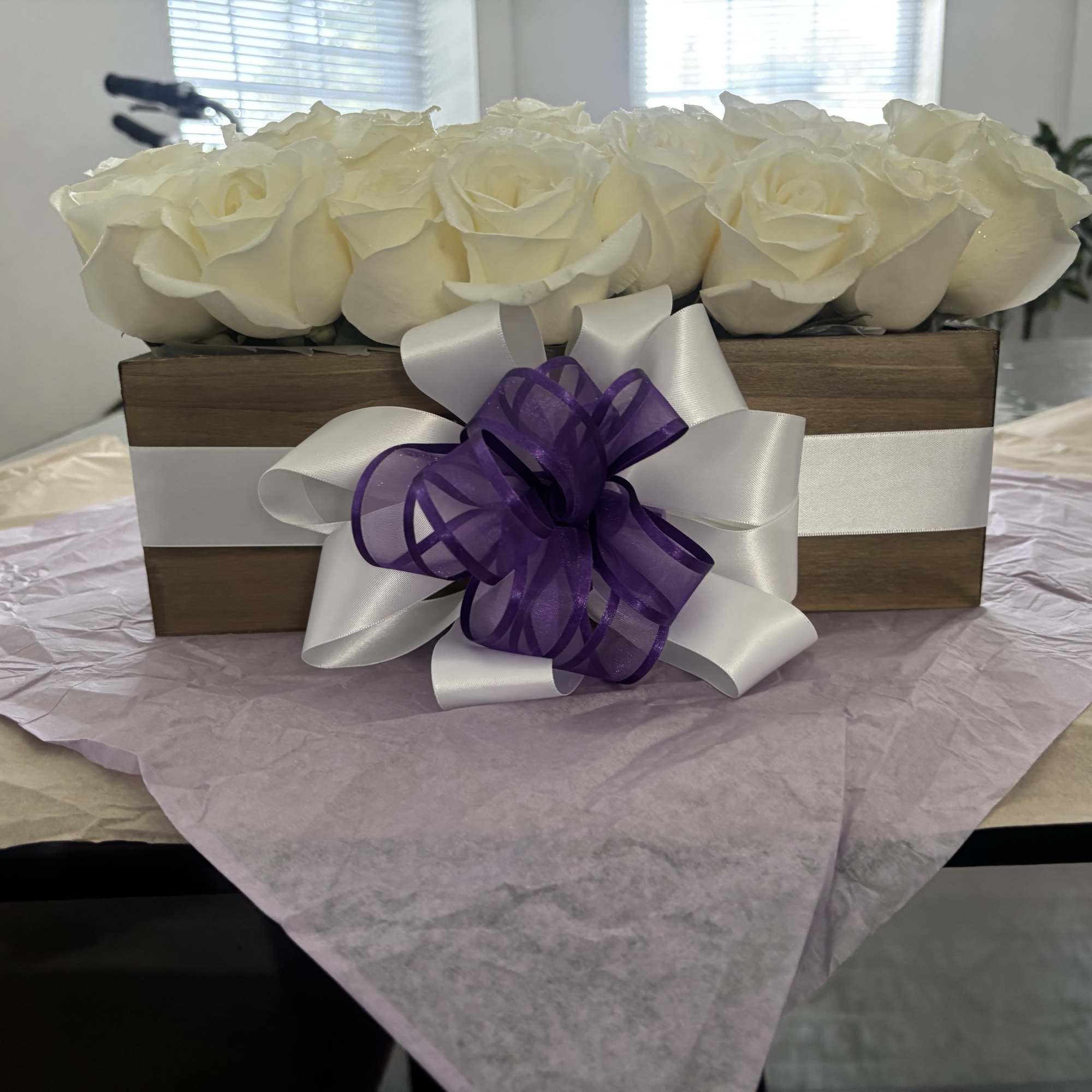 White roses arranged in a wooden box with a purple and white ribbon bow