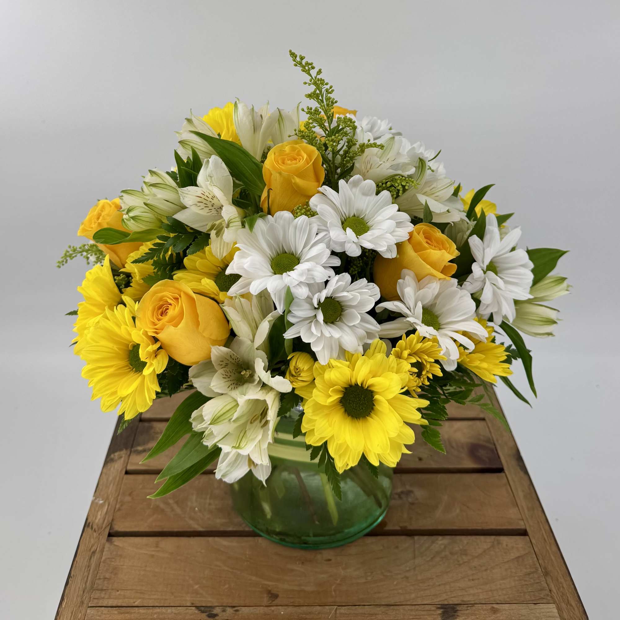 Yellow roses, white daisies, and white alstroemeria in a round green glass vase on a wooden crate