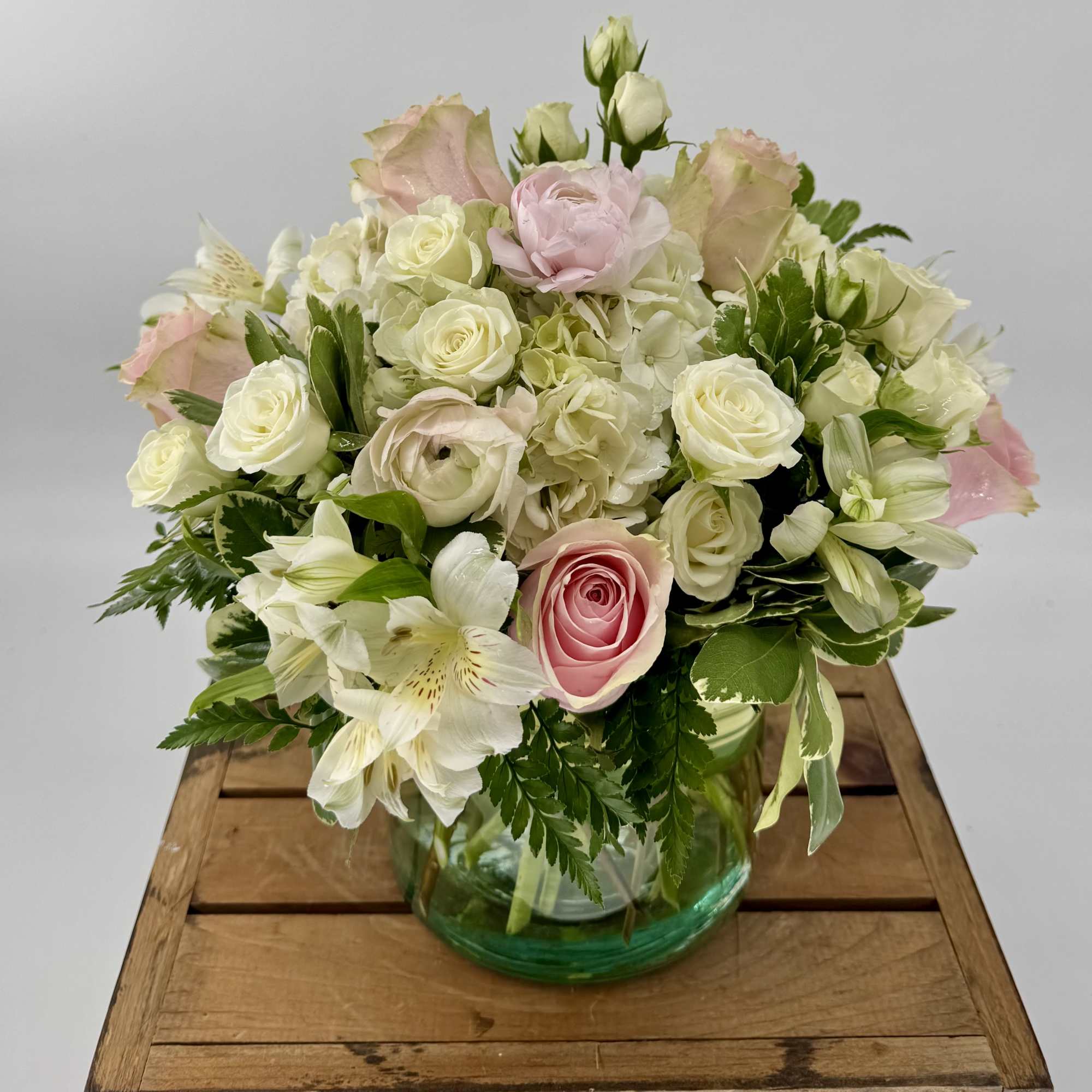 Round arrangement of white and pale pink roses, hydrangeas, and lilies in a glass vase