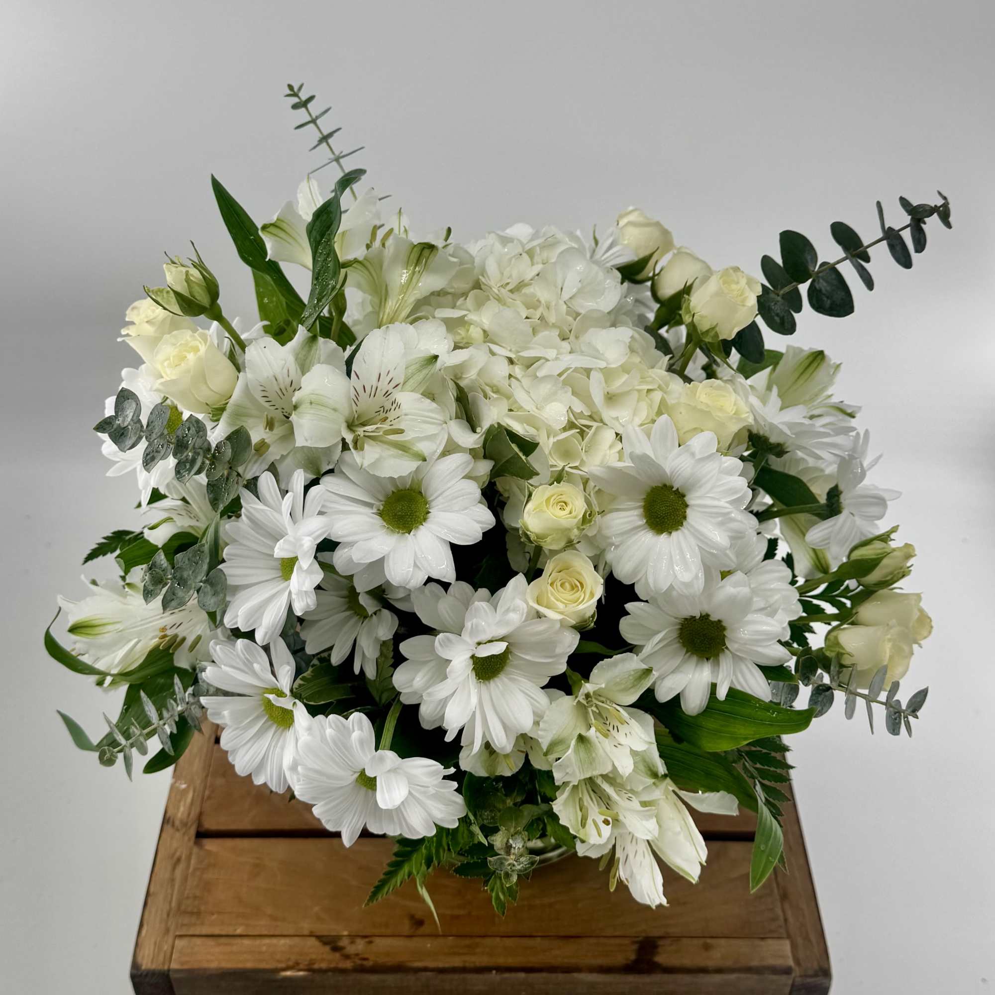 Round arrangement of white daisies, roses, and hydrangea in a low vase on a wooden crate