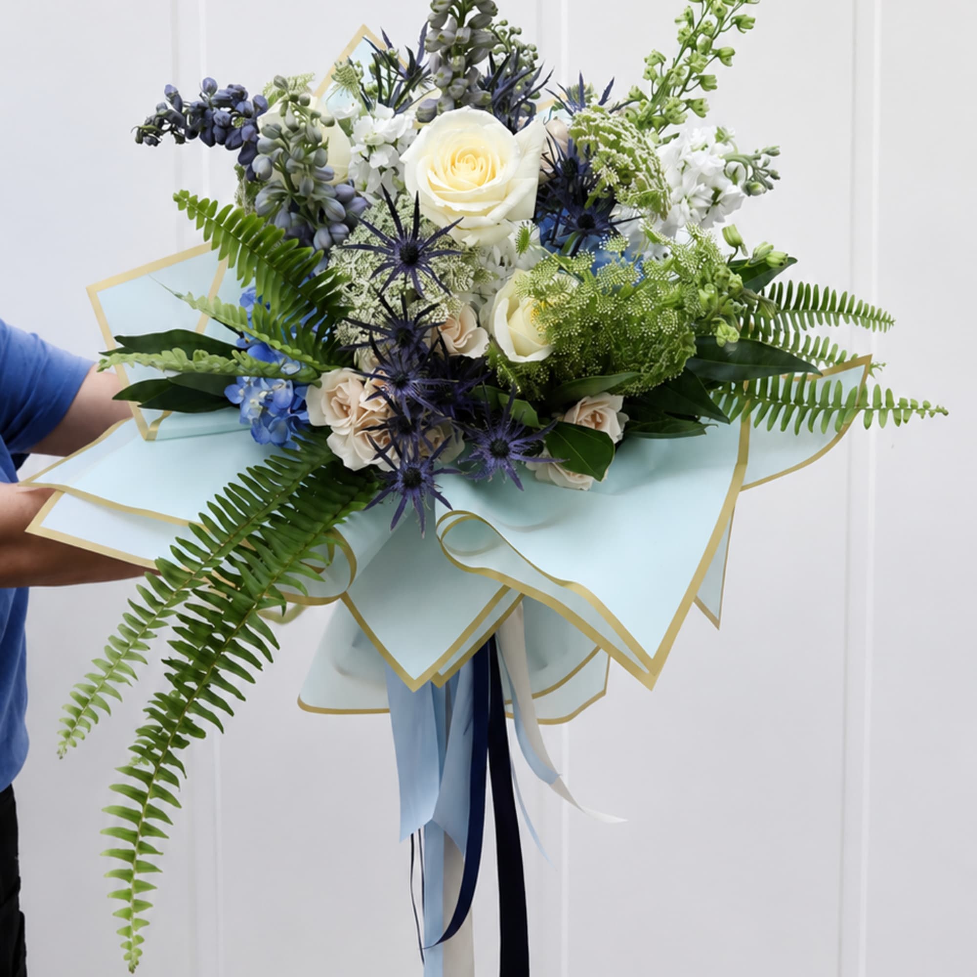 Handheld bouquet of white roses, blue flowers, and fern fronds wrapped in pale blue paper
