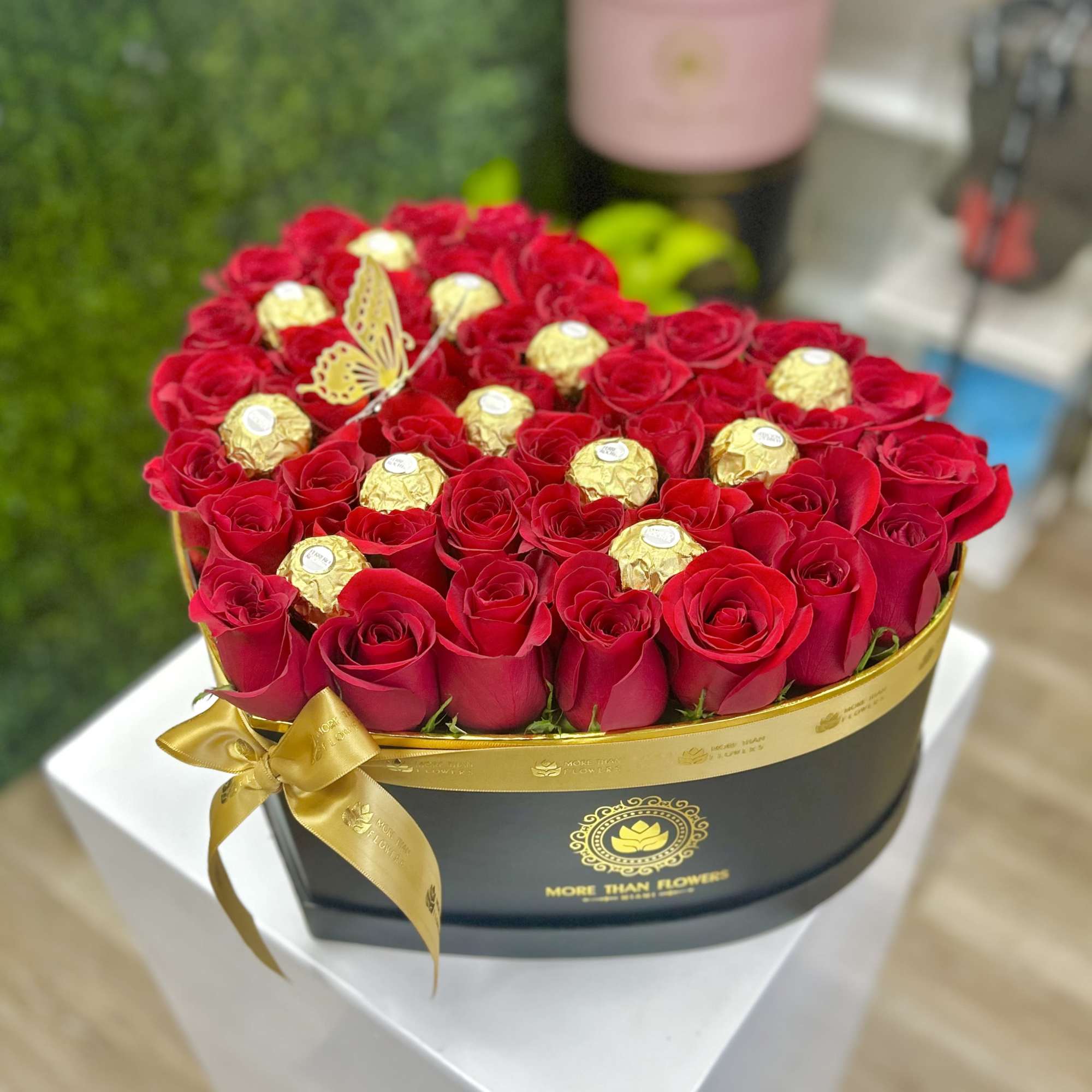 Heart-shaped black box of red roses with gold-wrapped chocolates and a gold ribbon bow.