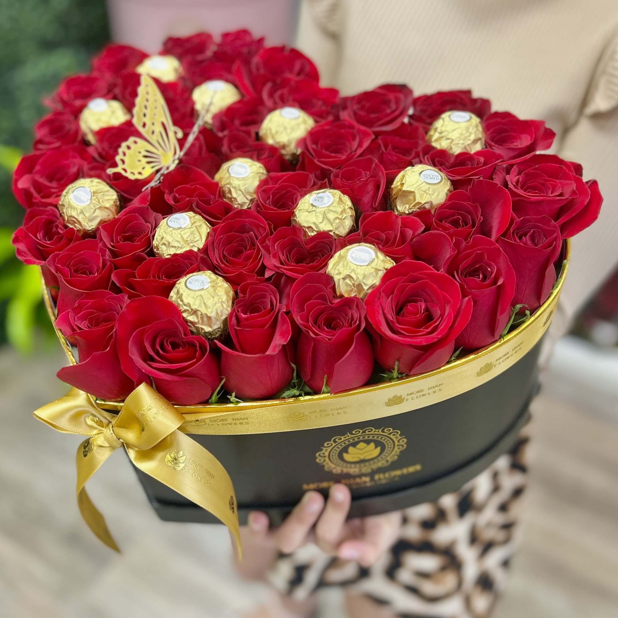 Heart-shaped black box filled with red roses and gold-wrapped chocolates, tied with a gold ribbon