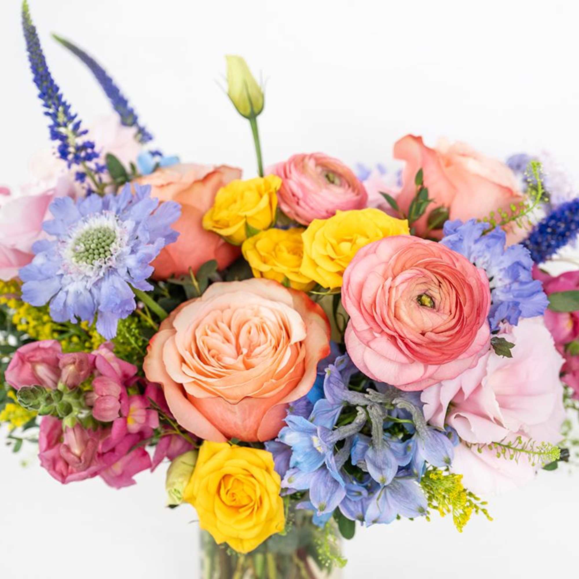 Bright mixed bouquet of peach roses, pink ranunculus, yellow roses, and blue flowers in a clear glass vase