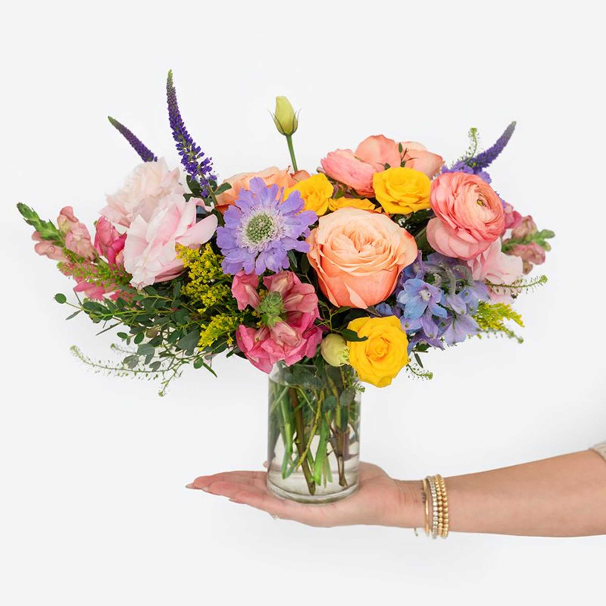 Colorful arrangement of roses, ranunculus, and mixed blooms in a clear glass vase held in one hand.
