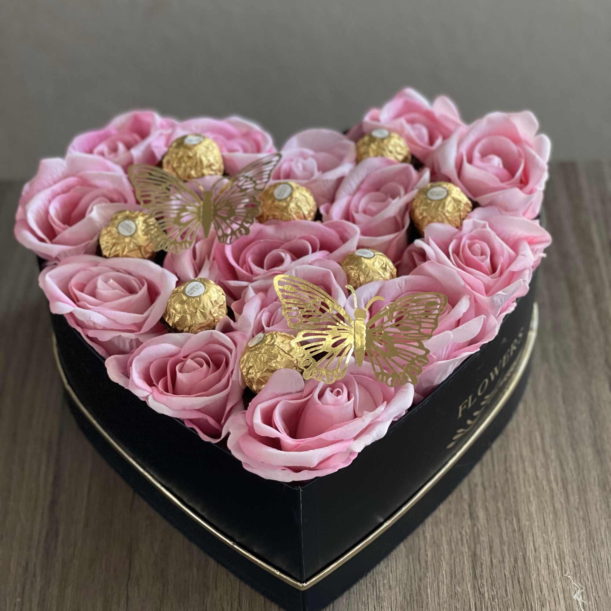 Pink roses in a heart-shaped black box with gold chocolates and butterfly picks