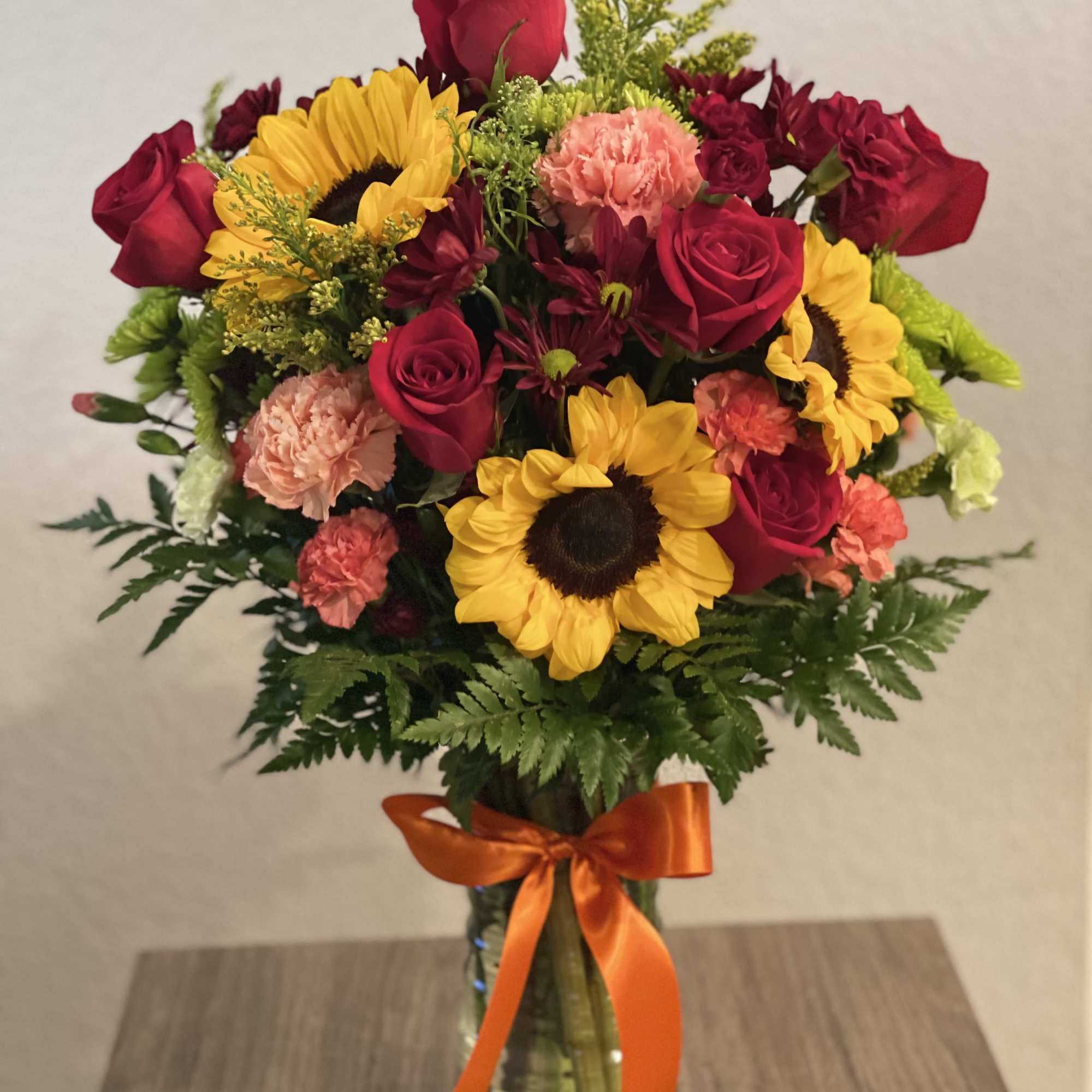 Bouquet of red roses, sunflowers, and pink carnations in a glass vase with an orange ribbon.