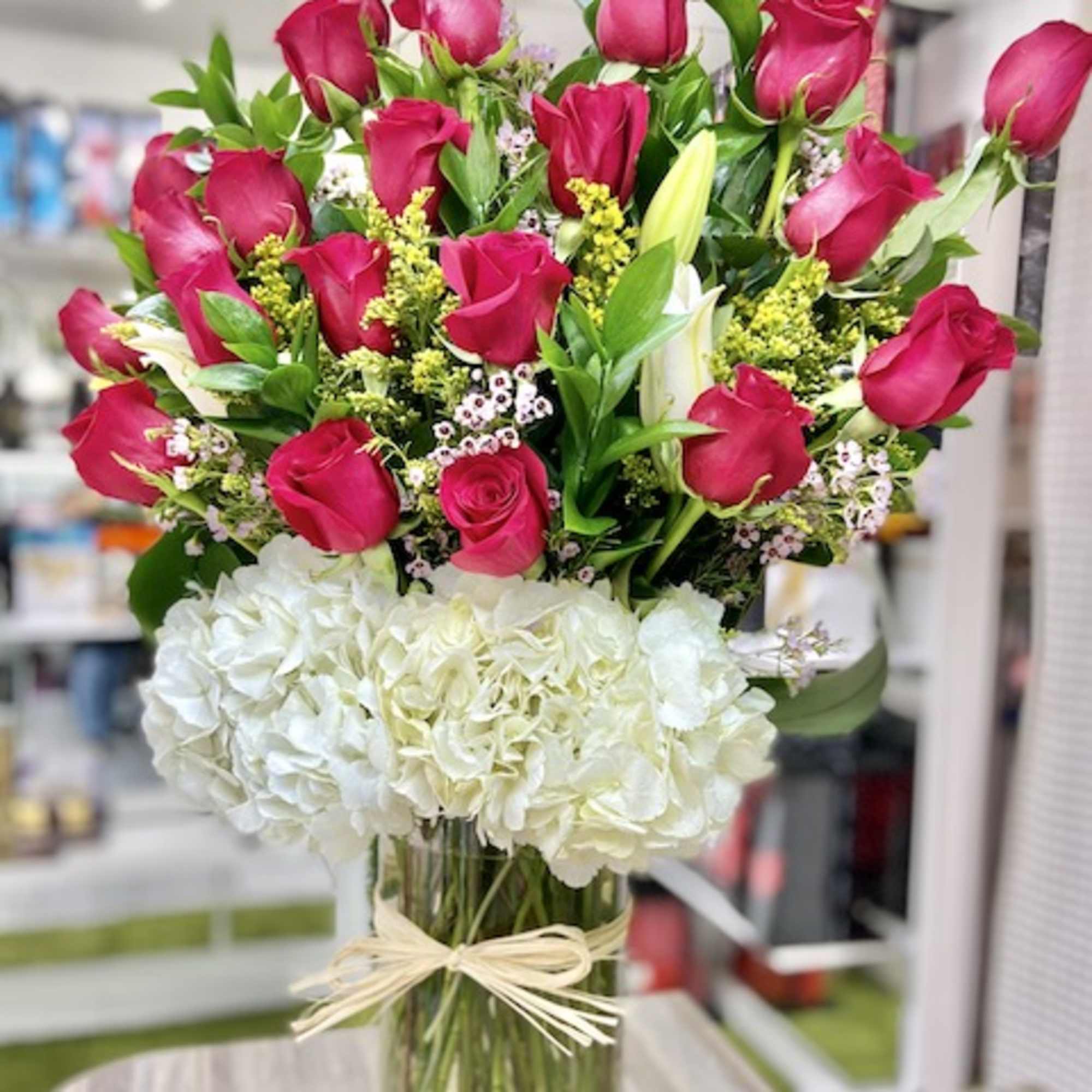 Tall arrangement of hot pink roses with white hydrangeas and lilies in a clear glass vase with raffia bow