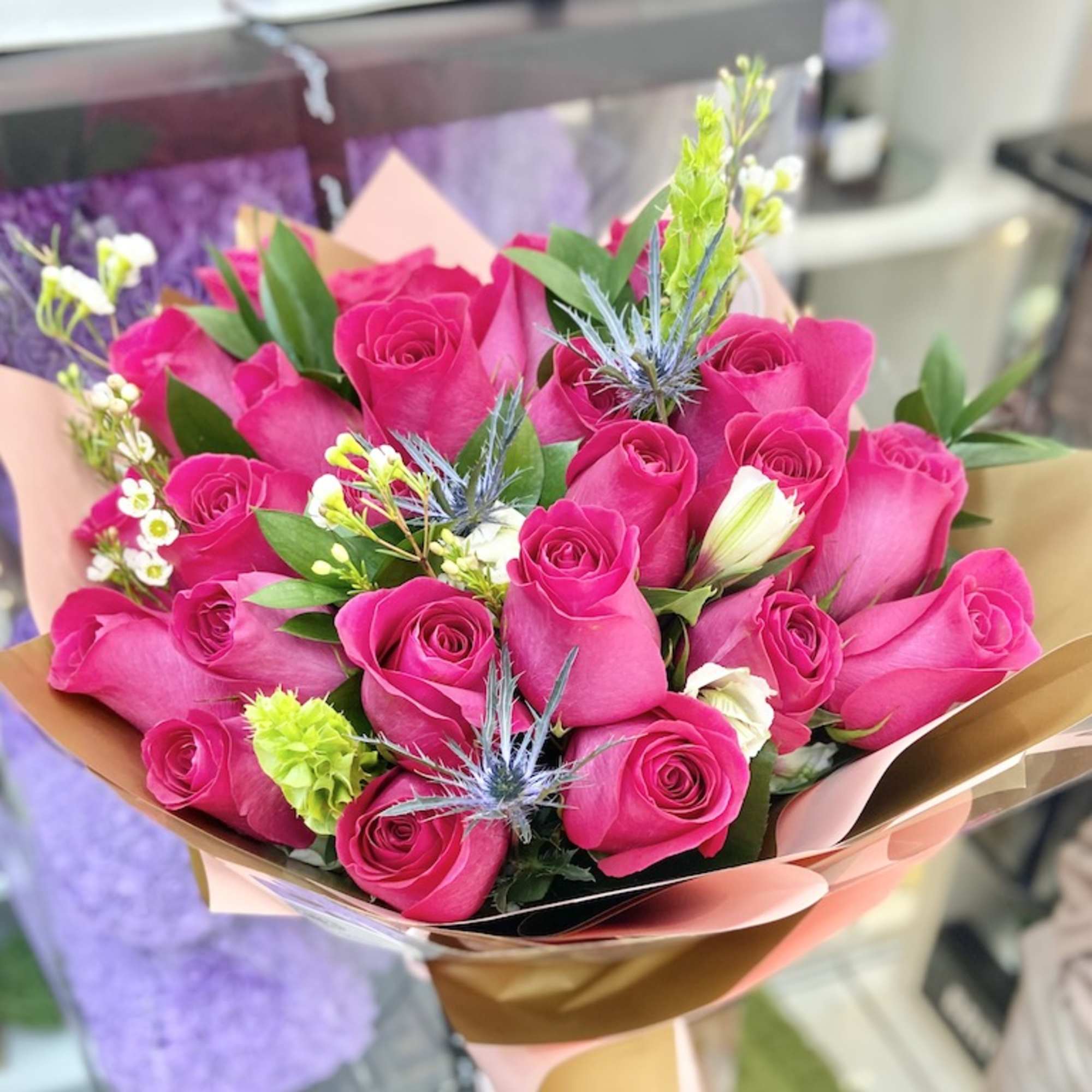 Hand-tied bouquet of bright pink roses with small white blooms and blue thistles wrapped in pink and gold paper