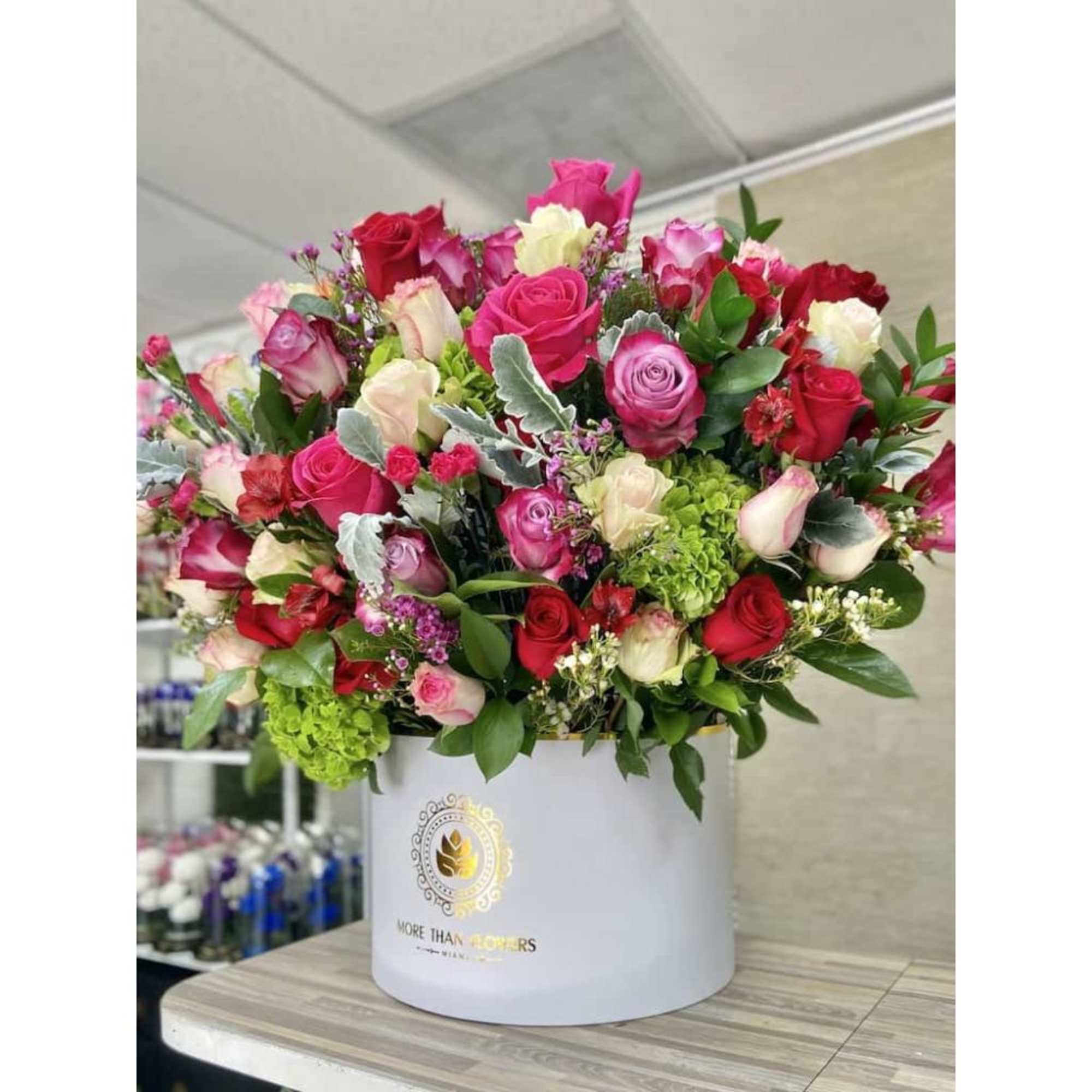 Large hatbox arrangement of red, pink, and cream roses with green hydrangea in a white container.