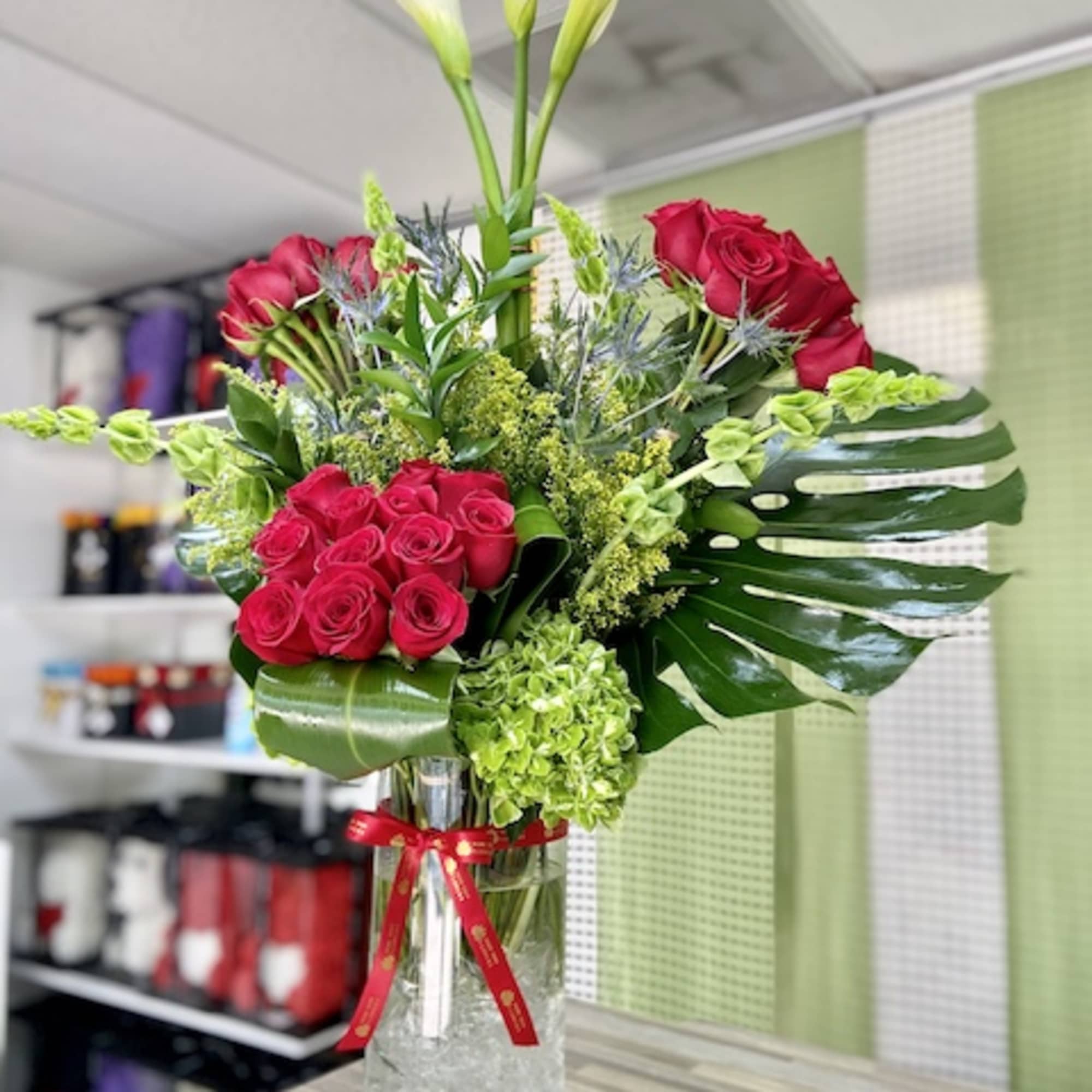 Tall arrangement of red roses, white calla lilies, and green hydrangea in a glass vase with a red ribbon.