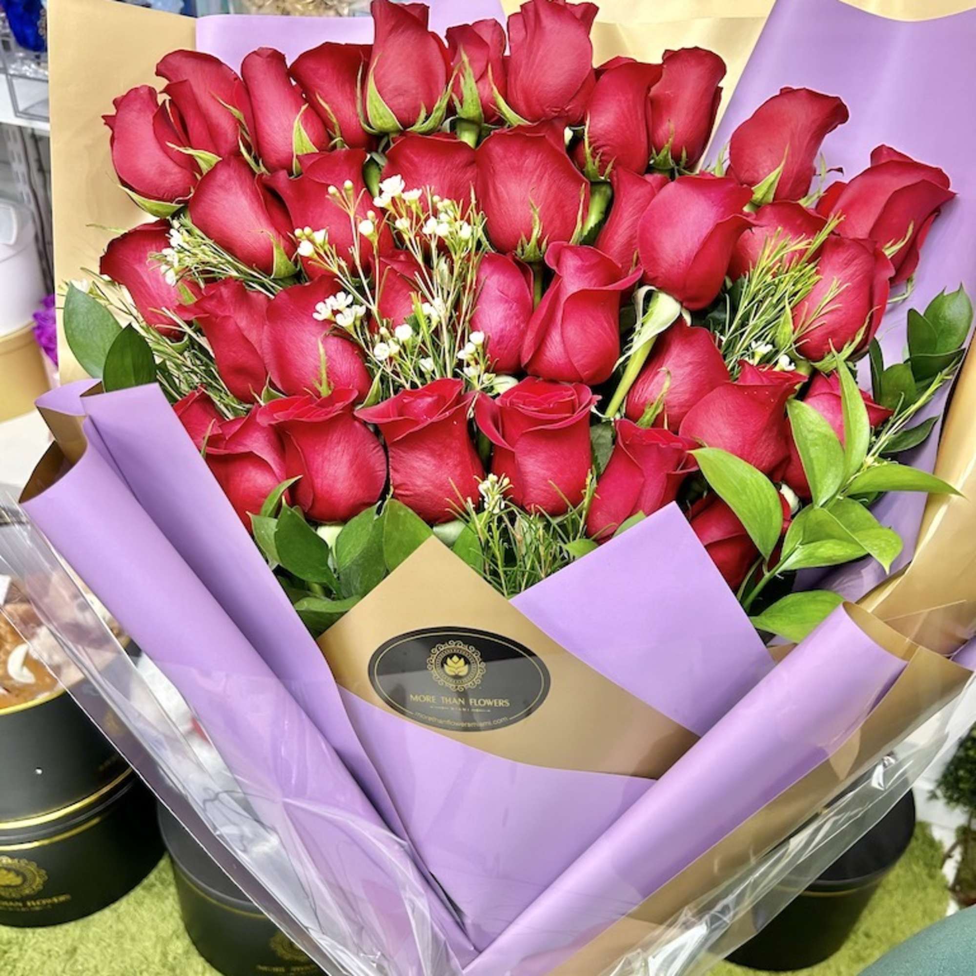 Large bouquet of red roses with small white blooms wrapped in tan and lavender paper with cellophane