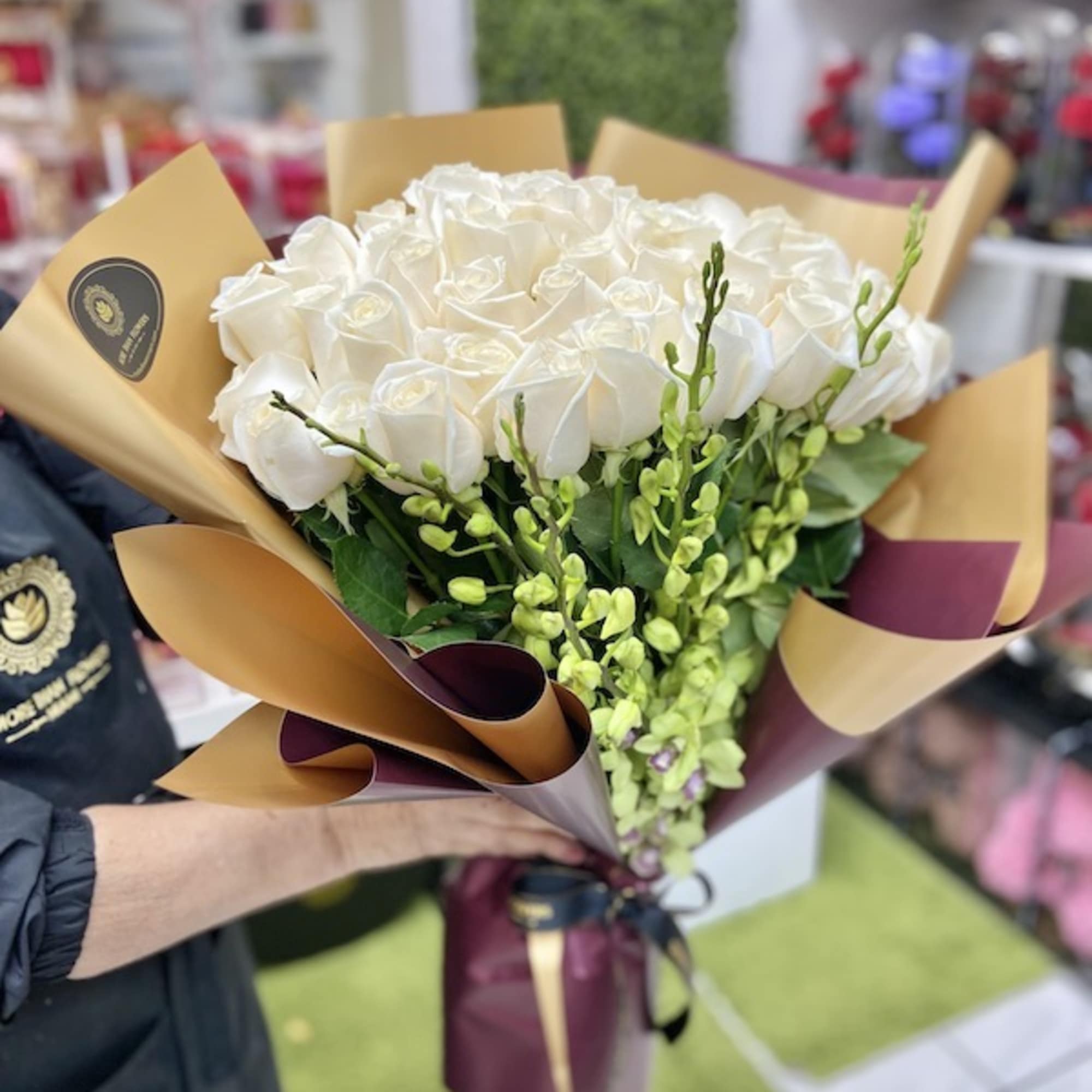 Large bouquet of white roses with green accent flowers wrapped in tan and burgundy paper.
