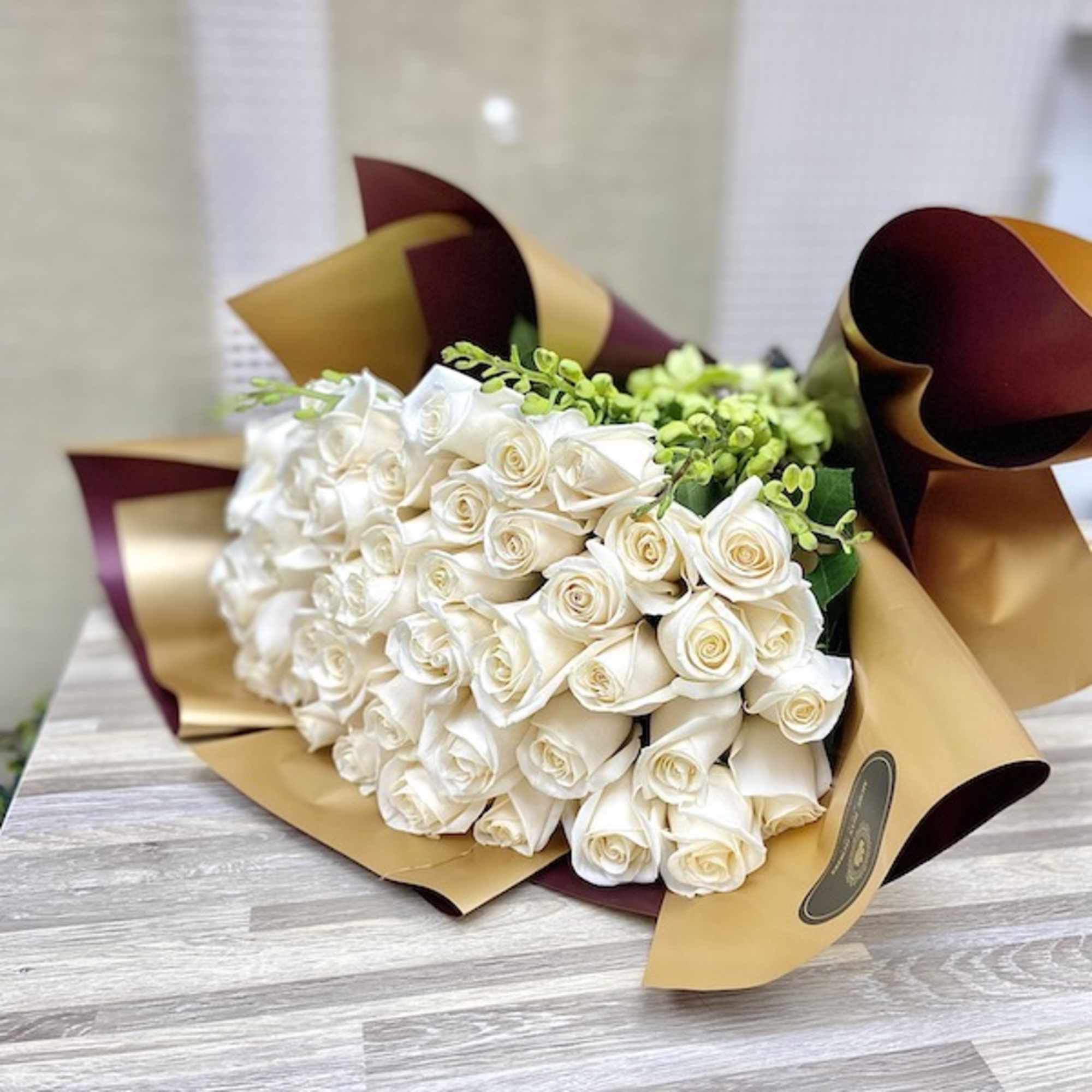 Large bouquet of white roses wrapped in gold and burgundy paper on a wooden surface