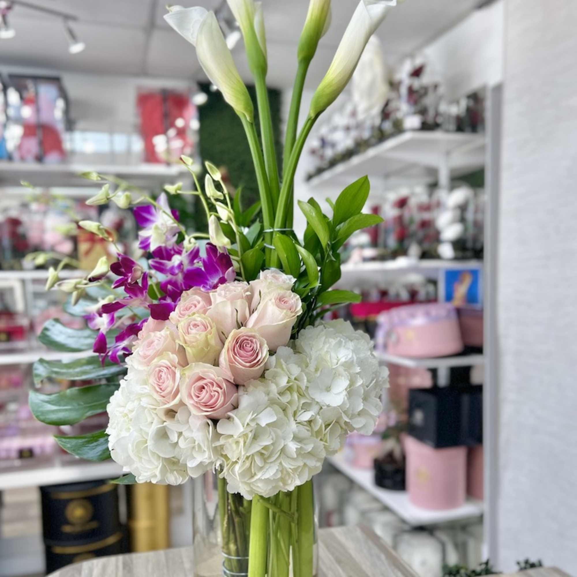 Tall arrangement of white calla lilies, hydrangeas, pink roses, and purple orchids in a clear glass cylinder vase
