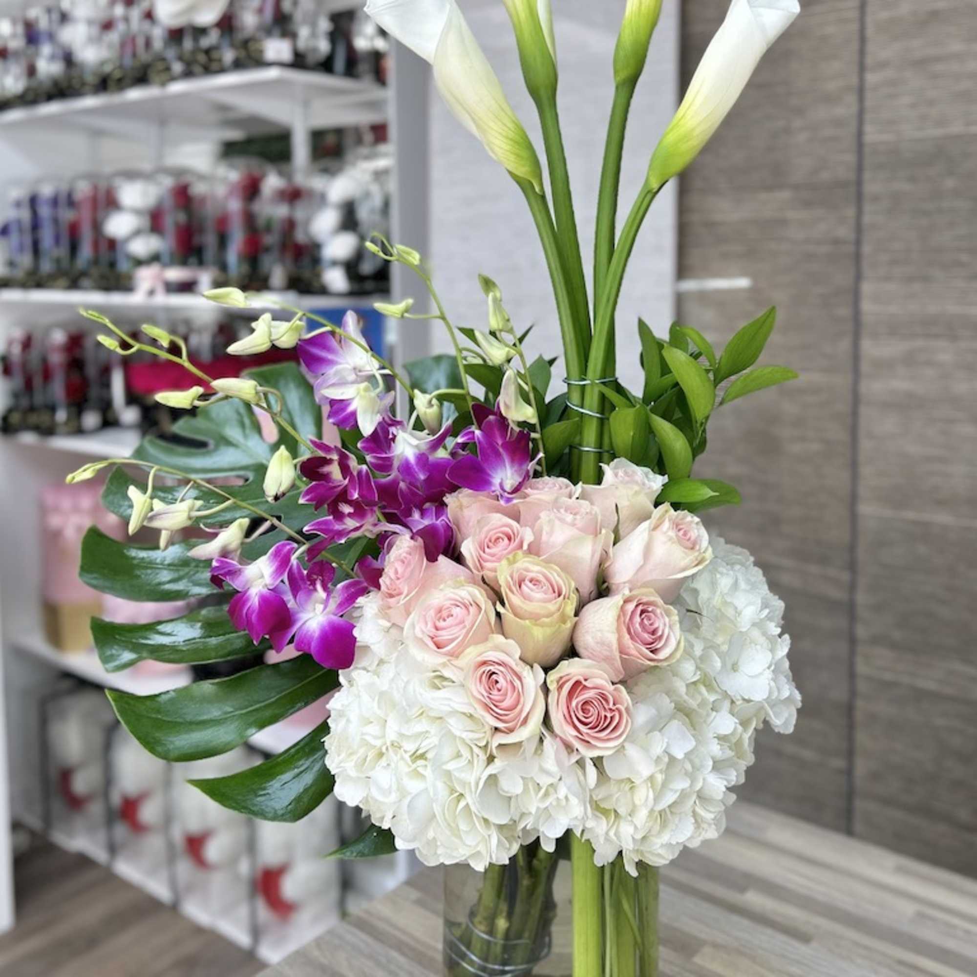 Tall arrangement with white calla lilies, pink roses, orchids, and hydrangeas in a glass vase with stones
