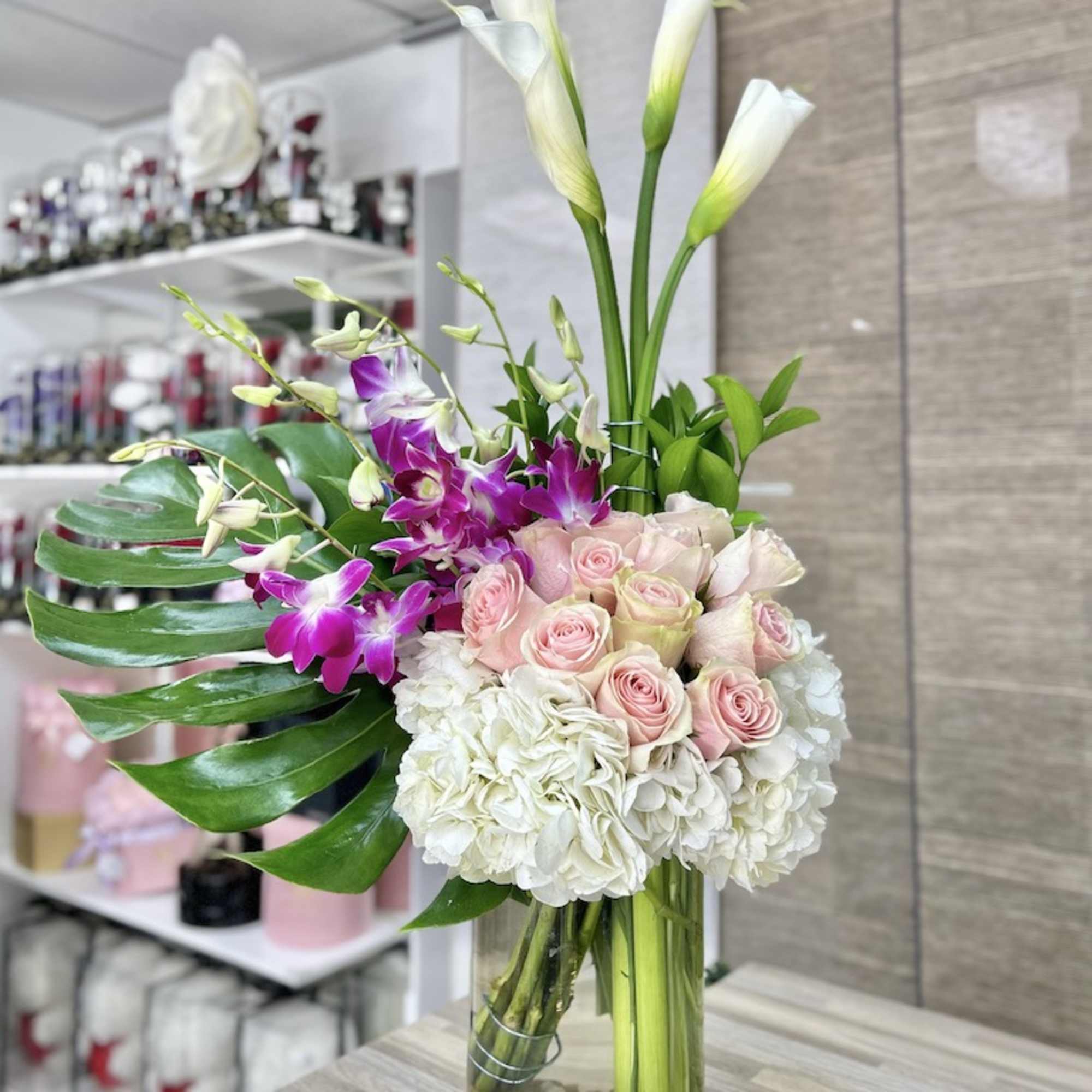 Tall mixed arrangement with white hydrangeas, pink roses, purple orchids and calla lilies in a clear glass vase