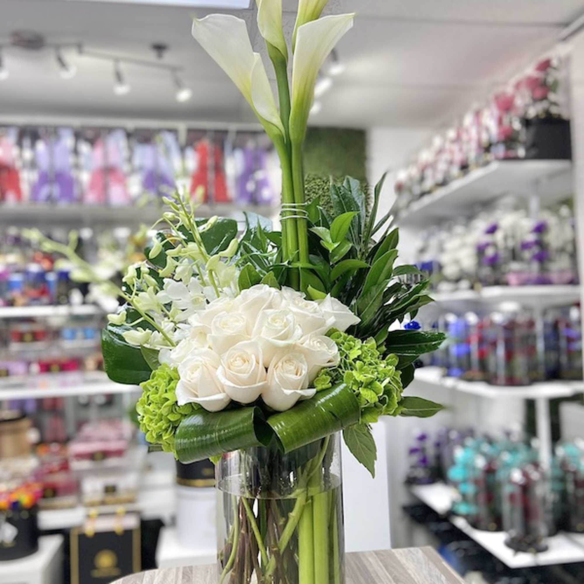 Tall white calla lilies and white roses with green accents arranged in a clear glass cylinder vase.