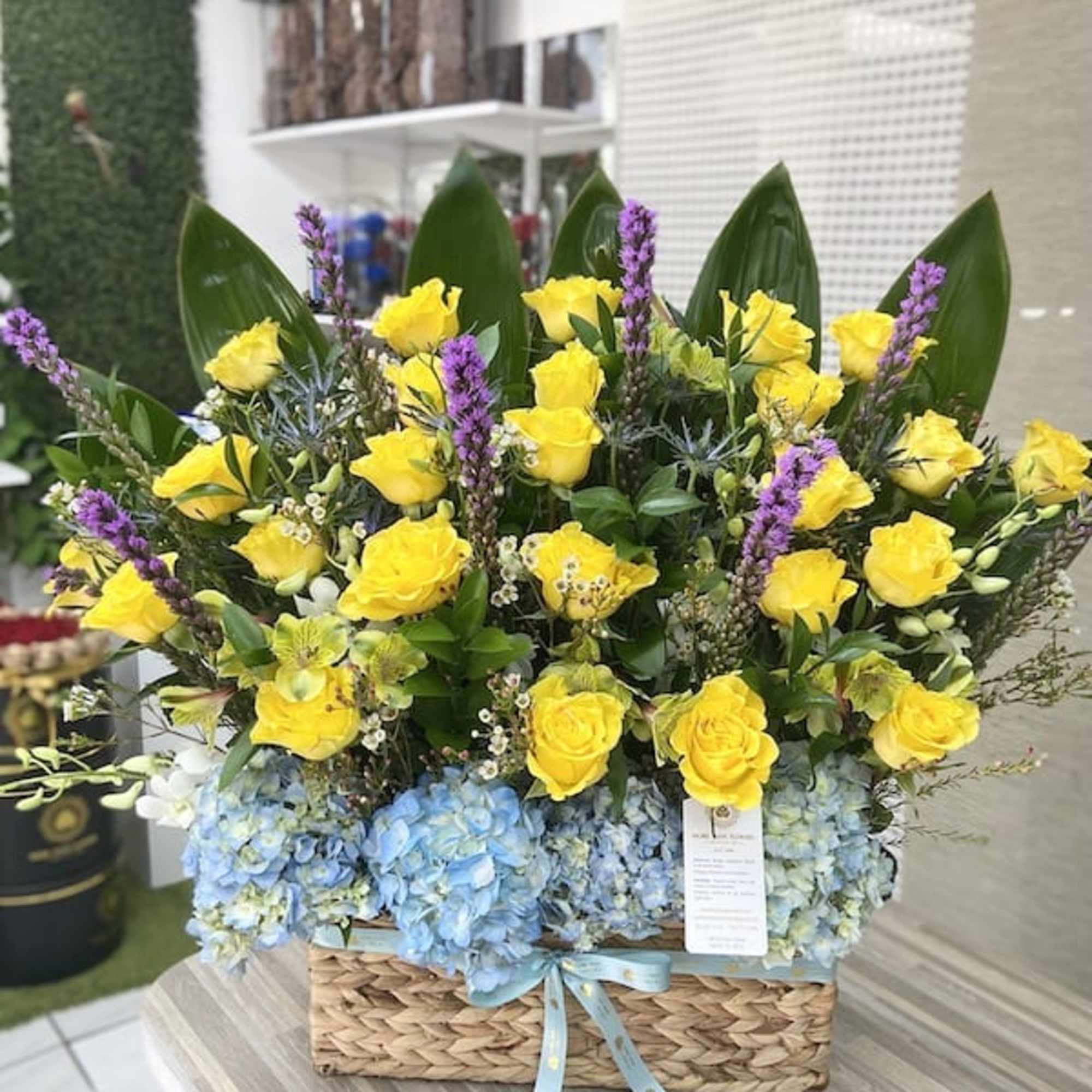 Tall basket arrangement of yellow roses and blue hydrangeas with purple flower spikes and a blue ribbon bow